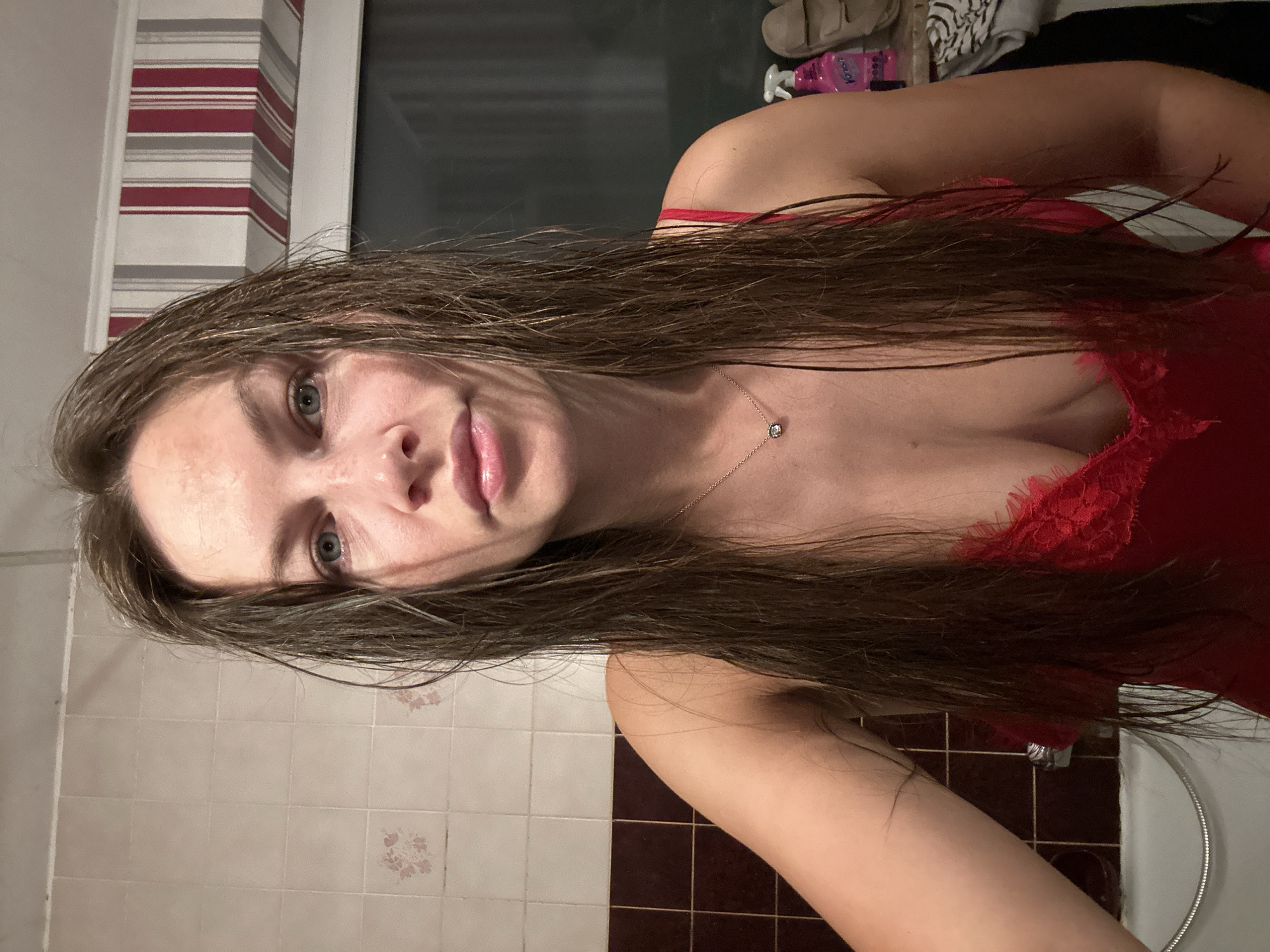 A front-facing portrait of an adult female with long, wet, dark brown hair, captured in a bathroom setting in Fredenbeck, Germany. The woman is looking directly into the camera with a neutral expression, her light-colored eyes prominent. She is wearing a red, lacy camisole with thin straps and a delicate necklace featuring a small, dark-colored pendant. Her wet hair frames her face and extends over her shoulders and chest.
The background consists of tiled walls: a lighter, possibly off-white, upper section with faint floral patterns, and a darker brown lower section. A white bathtub or shower basin and a metallic flexible hose are visible on the lower left. On the right, a window or dark glass pane is partially visible, topped by a valance with red, white, and gray vertical stripes. A shelf or counter to the right holds a pink spray bottle, two light-brown shoes/slippers, and a zebra-patterned towel.