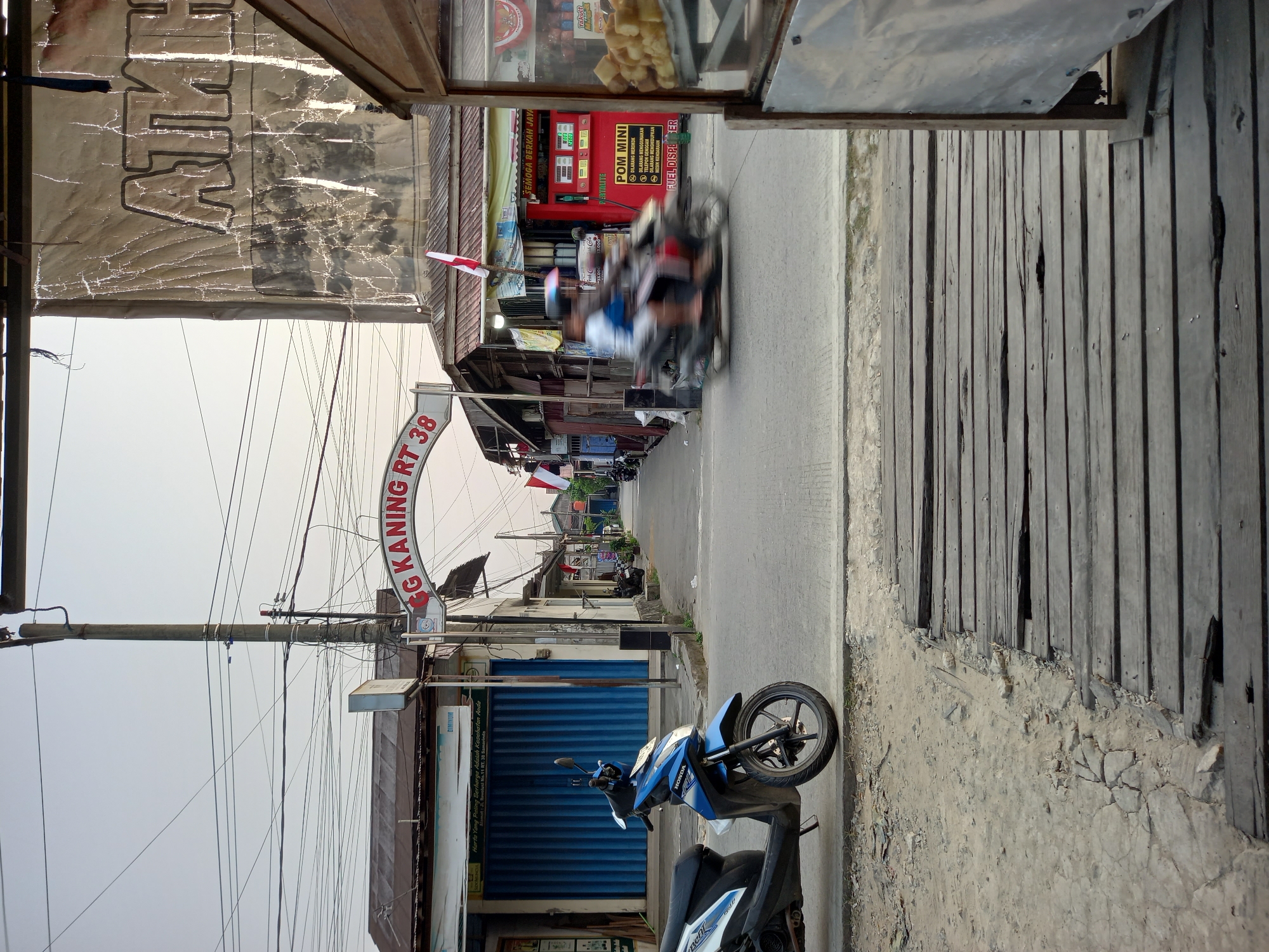 The image depicts a street scene in Samarinda, Indonesia, likely in a residential or commercial area given the shops and the street sign. A blue and black Honda scooter is parked on the left side of the foreground, facing the viewer. Beyond the scooter, the street extends into the distance, lined with buildings. A prominent arched sign over the street reads "GG KANING RT 38", indicating the street name and its residential zone. On the right side of the street, a small gas station with red pumps is visible, labeled "POM MINI". Two figures on a motorbike are captured in motion, blurred due to their speed, passing the gas station. In the foreground, a weathered wooden walkway or bridge made of planks is present, with some sections broken and uneven. Overhead, a complex network of electrical wires crisscrosses the sky. A large, somewhat tattered banner with faded text is mounted on a building on the right side of the image. The sky is overcast and light, suggesting either early morning or late afternoon.