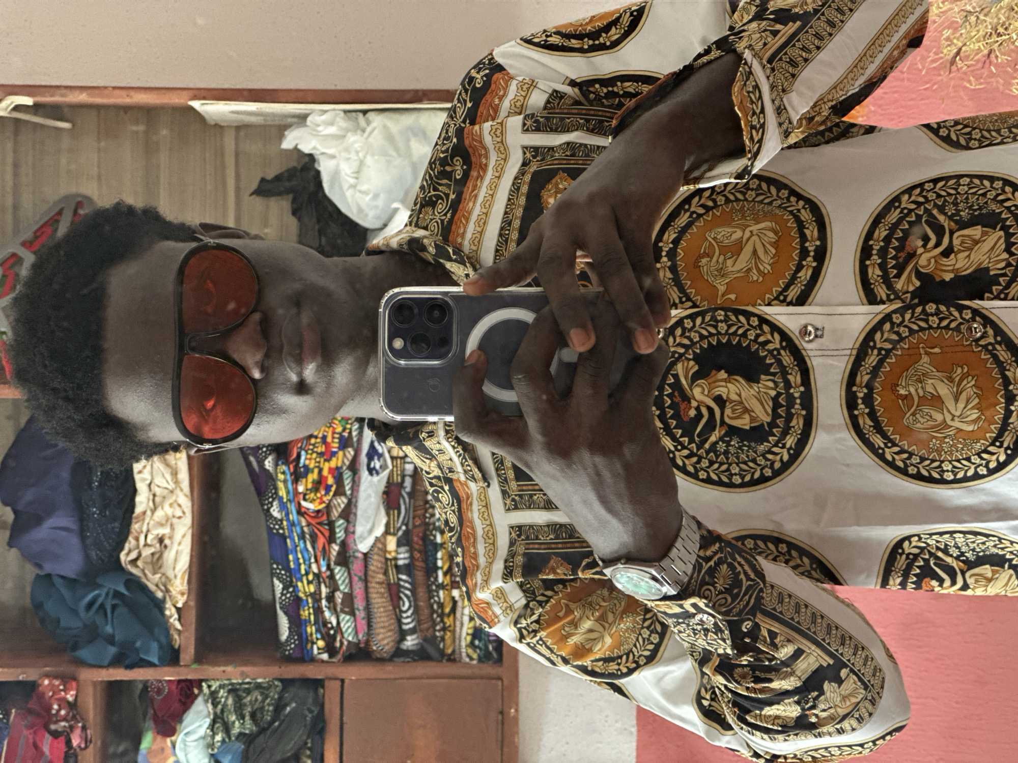 A medium shot depicts a man in sunglasses taking a selfie in Badagry, Nigeria. He is holding a smartphone, displaying his reflection. He wears a white shirt with ornate circular patterns in gold and black. A silver wristwatch adorns his left wrist. Behind him is a shelf containing folded textiles of various colors and patterns. The backdrop features pale brown and pink wall colors.