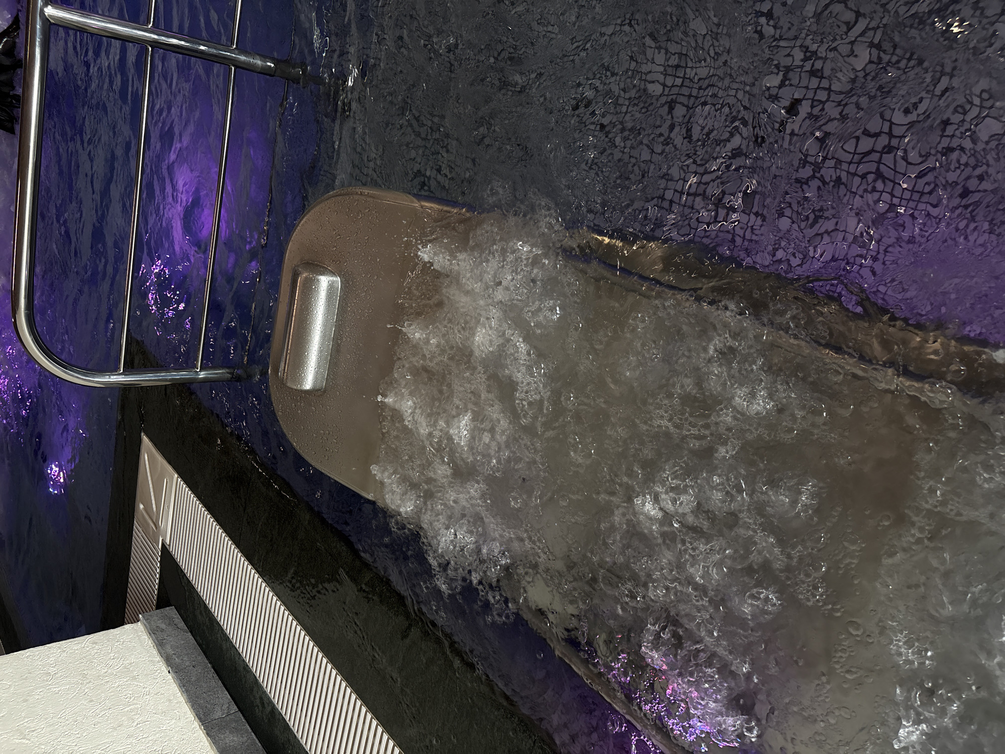 An overhead shot shows a section of a swimming pool with bubbling water. A metallic bar with horizontal rails crosses the frame diagonally from the upper right to the lower left. In the foreground, a tan-colored submerged lounger with a metallic headrest is producing jets of water that create a foamy, agitated surface. The pool water is illuminated with purple lighting, casting reflections on the surface and highlighting the tiled bottom. To the left of the lounger, a drainage grate with parallel metal slats runs along the edge of the pool, bordering a white tiled wall. The scene is devoid of people. The location is not determinable from the image.