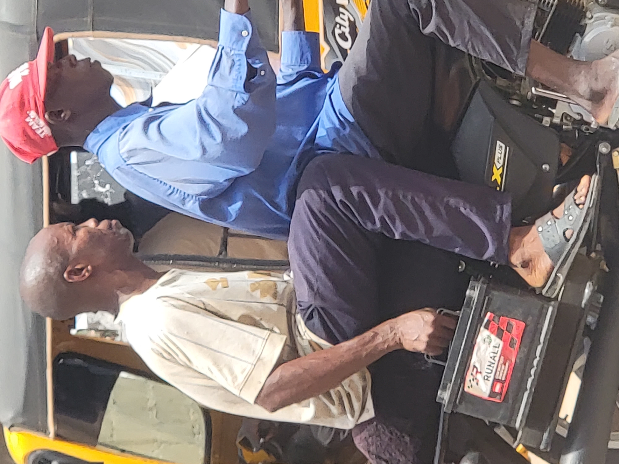 Two adult males are seated in an auto rickshaw, facing opposite directions. The man on the left wears a short-sleeved, light-colored shirt. The man on the right wears a red baseball cap and a light blue long-sleeved shirt. Both men wear dark trousers. The rickshaw’s engine is visible. A battery with "RUNALL" printed on it rests near the bottom right. Based on the presence of auto rickshaws, the image appears to be taken in a urban location, potentially Maiburiji, Nigeria.