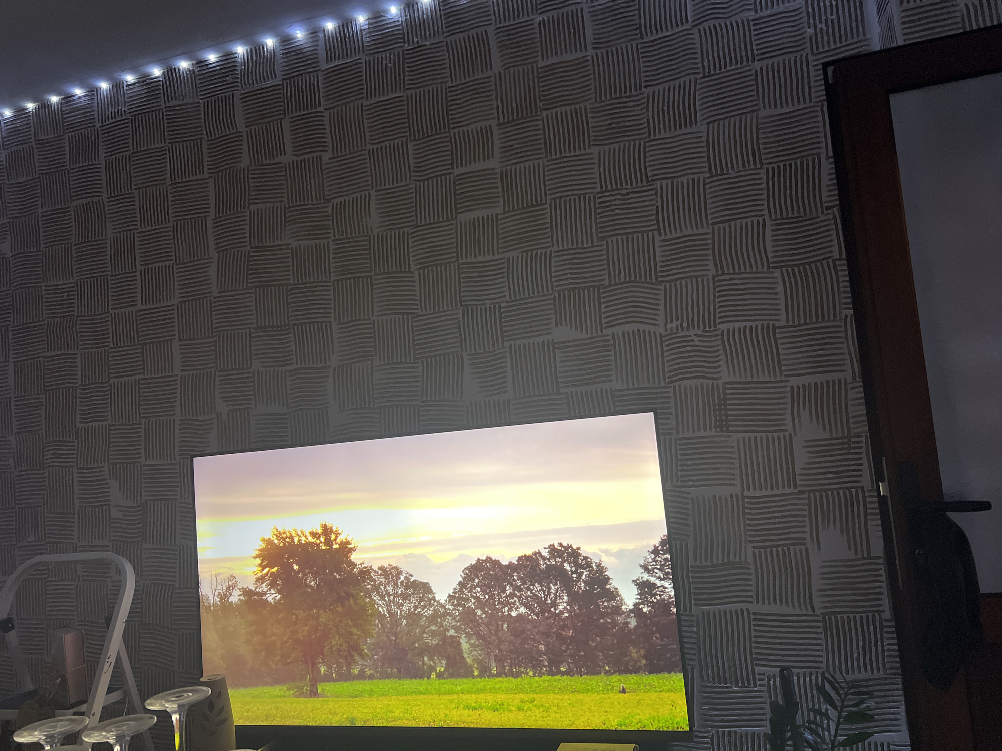 In Medkovets, Bulgaria, a living room scene is captured. The room features a patterned wallpaper with a grid of square motifs, each filled with parallel lines. A string of white LED lights runs along the top of the wall. Dominating the lower portion of the frame is a large television screen displaying a vibrant landscape scene. The screen depicts a sunset with a bright yellow and orange sky, casting a warm glow over a lush green field and silhouetted trees. To the left of the television, a white stepladder and what appears to be a wine decanter are visible. On the right side of the image, a dark wooden doorframe is present, with a partial view of a dark interior.
