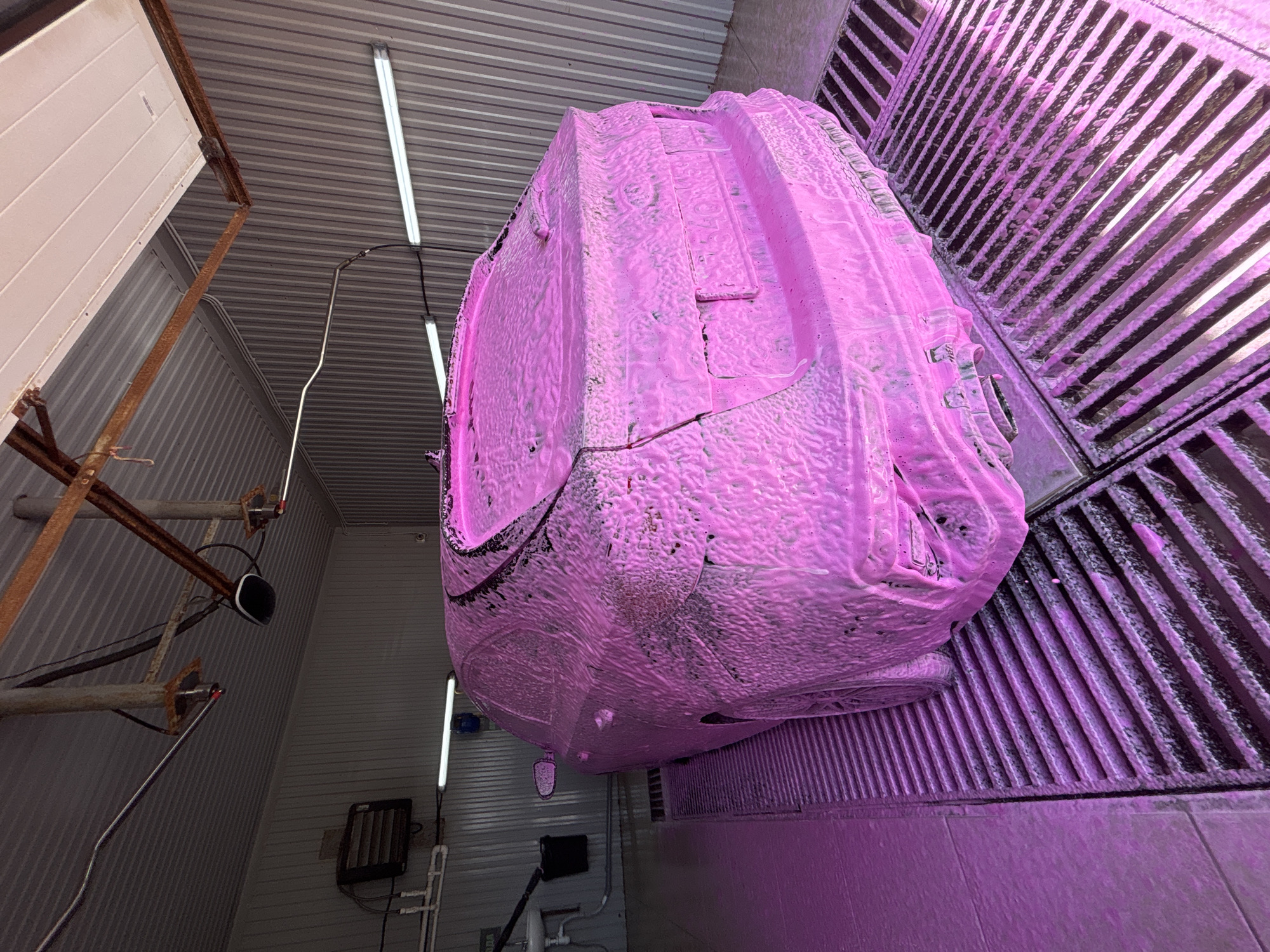 The image depicts a vehicle, heavily coated in pink foam, positioned over a metal grate within an enclosed structure. The structure's walls are corrugated metal, illuminated by linear fluorescent lights. Rusty metal supports are visible on the left wall. The scene is likely a car wash facility. The presence of the name 'Chaikovsky, Russia' appears to be extraneous to the image content. No people are visible.