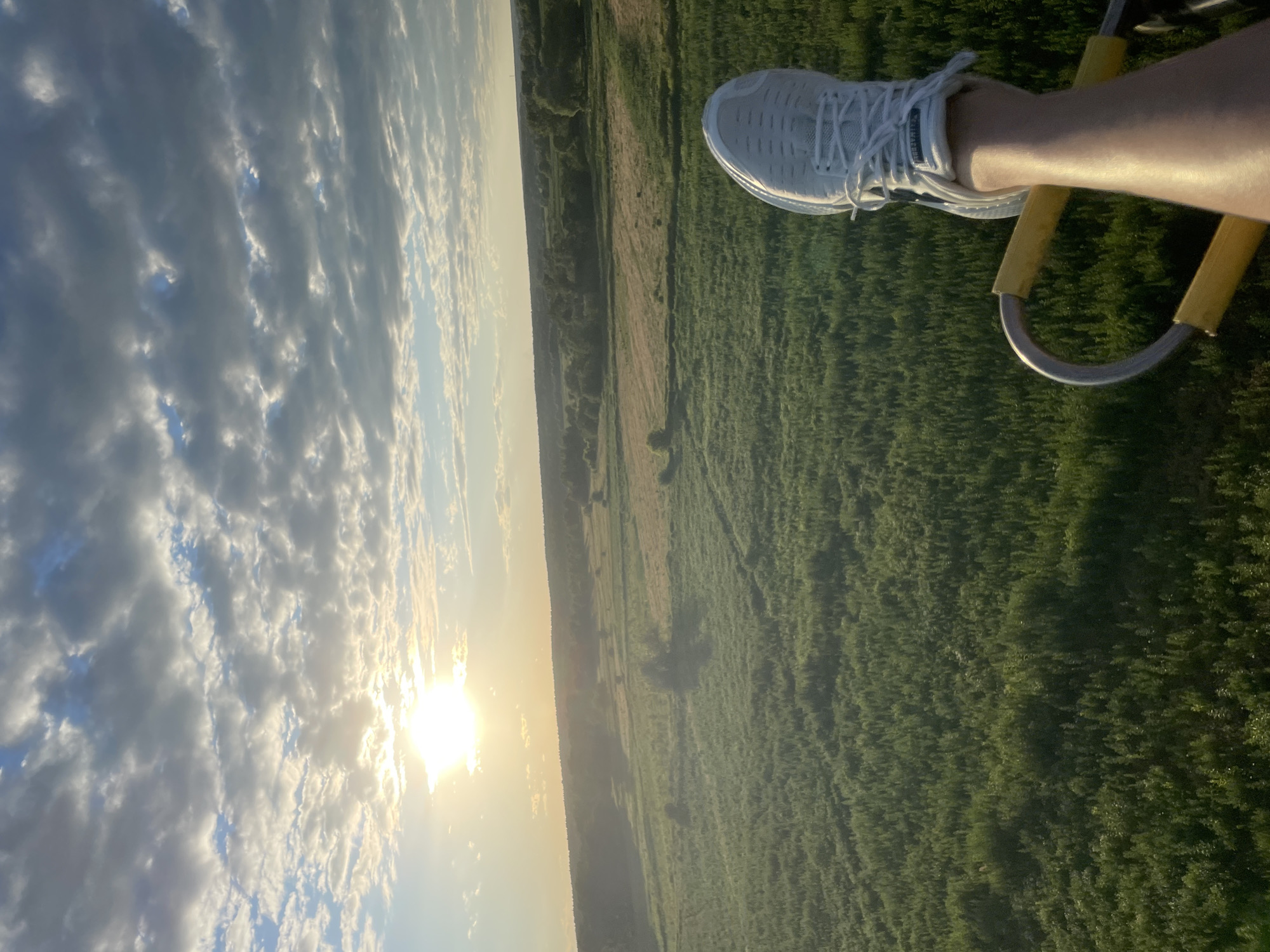 The image captures a first-person perspective from a hot air balloon ascending over a rural landscape. In the foreground, a person's right leg and foot, clad in a white athletic shoe, are visible, resting on a yellow handle of the balloon's basket. The leg is illuminated by sunlight, suggesting a warm day. The background reveals a sprawling green field dotted with trees and some patches of drier vegetation, indicative of farmland. A distant tree line forms the horizon under a sky filled with a mixture of white and grey clouds. The sun is partially obscured by the clouds, casting a warm, golden light across the scene. The geographical location is specified as Турово, Russia.