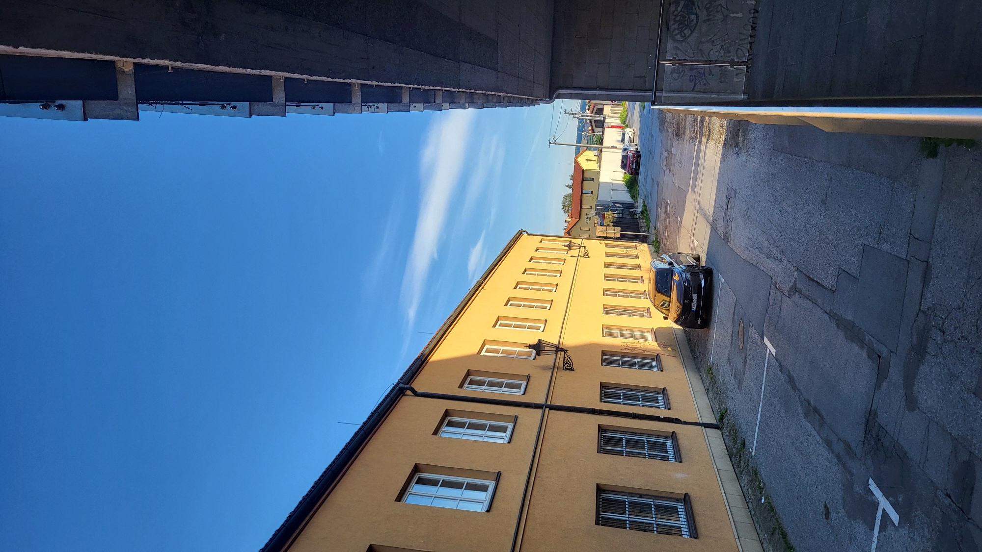 The image depicts an urban scene with a narrow street flanked by buildings. On the left, a two-story building with a yellow facade features numerous white-framed windows, some with security bars. A vintage-style street lamp is mounted on the wall. The street itself is paved with asphalt and shows signs of wear, with parking lines visible. A dark-colored SUV is parked on the right side of the street. Further down the street, other vehicles are visible, and on the right side of the frame, a modern concrete structure with exposed beams and a railing suggests a new construction or elevated walkway. The sky is a clear blue with a few wispy clouds. The lighting indicates it is likely late afternoon or early morning, with the sun casting long shadows.
The specific location is believed to be on or near Južná trieda street in Prešov, Slovakia. This street is known for its mix of older residential and commercial buildings, as well as newer construction projects, which aligns with the visual elements present in the photograph. The presence of parked cars and street lamps further supports an urban environment like Prešov.