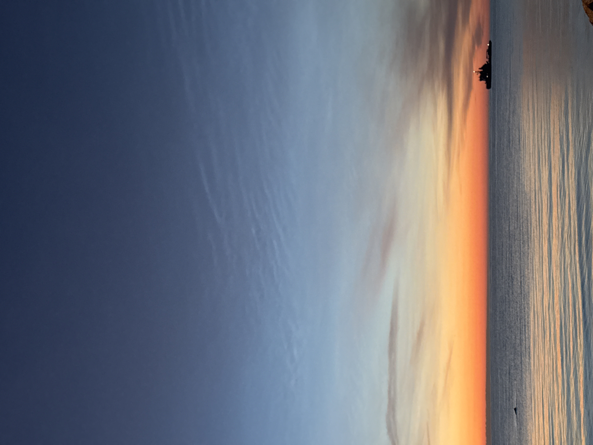 A photograph captures a serene ocean sunset with a silhouette of a ship on the horizon. The sky displays a gradient of colors, transitioning from a deep blue at the top to warm oranges and pinks near the horizon. The calm sea reflects the sky's colors, creating gentle ripples on the surface. In the distance, a ship with a lit superstructure is visible. To the left of the ship, a small dark shape, possibly a marine animal, breaks the water's surface. The scene appears to be a quiet evening over the sea, with no discernible human presence in the immediate vicinity. Given the presence of a ship and open water, this scene could be depicted off the coast of Tallinn, Estonia, a city known for its maritime activity and scenic Baltic Sea views.