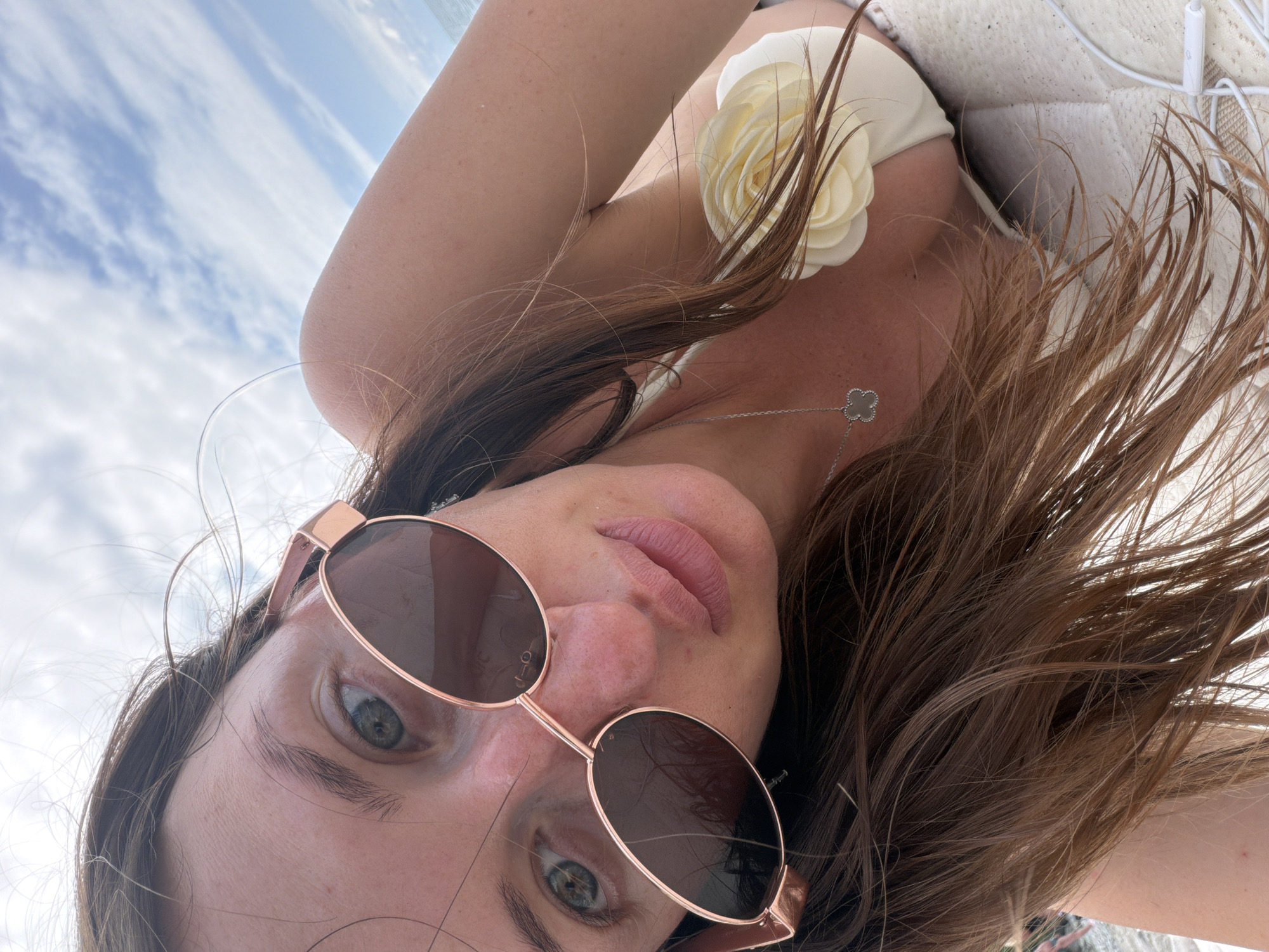 A woman is lying on a beach towel, her head tilted upwards and slightly to the right, looking at the camera. She is wearing a white bikini top with a floral embellishment. Her face is partially obscured by large, rose-gold tinted sunglasses with oval lenses. Her long, brown hair is spread around her head and shoulders. The background shows a cloudy sky and the distant sea. The image appears to have been taken in Tsikhisdziri, Georgia, likely during daylight hours.