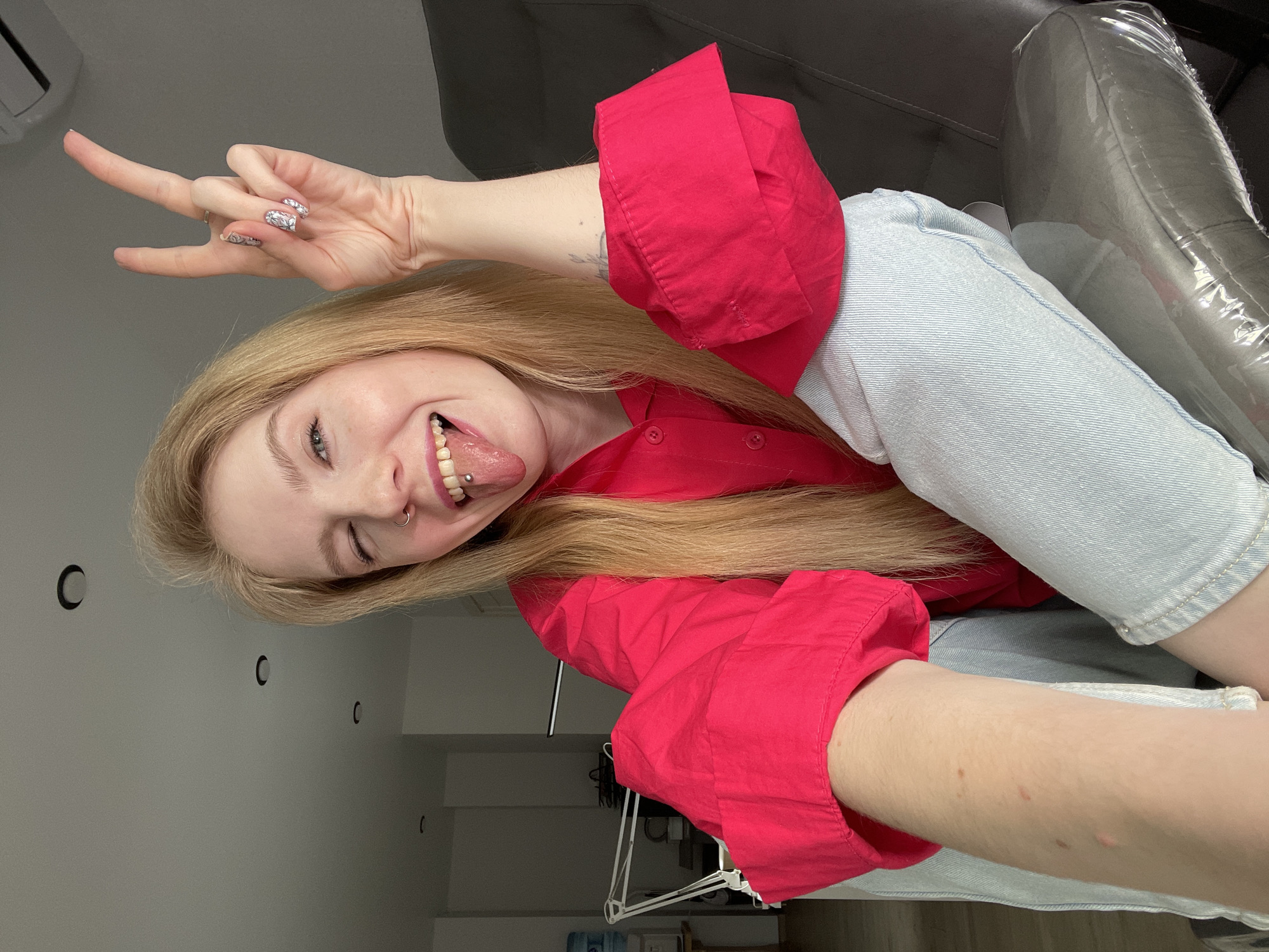 A young blonde woman is featured in a playful, vertically oriented selfie, captured from a slightly low angle. She is winking, sticking out her tongue (which has a barbell piercing), and making a "rock on" hand gesture with her left hand. A small hoop nose ring is also visible. Her nails are adorned with intricate black and white floral patterns. She is wearing a vibrant red long-sleeved button-up shirt and light blue denim jeans. A faded tattoo can be seen on her right forearm.
The setting is an indoor room with light grey walls and several black recessed lights. An air conditioning unit is partially visible in the upper left. To the woman's right, a grey piece of furniture, possibly a chair or sofa, is covered in clear plastic wrap. Indistinct furniture or equipment, including a white metal stand, can be seen in the background. The woman's expression is cheerful and energetic. No visible text is present in the image. The location context is Izhevsk, Russia.