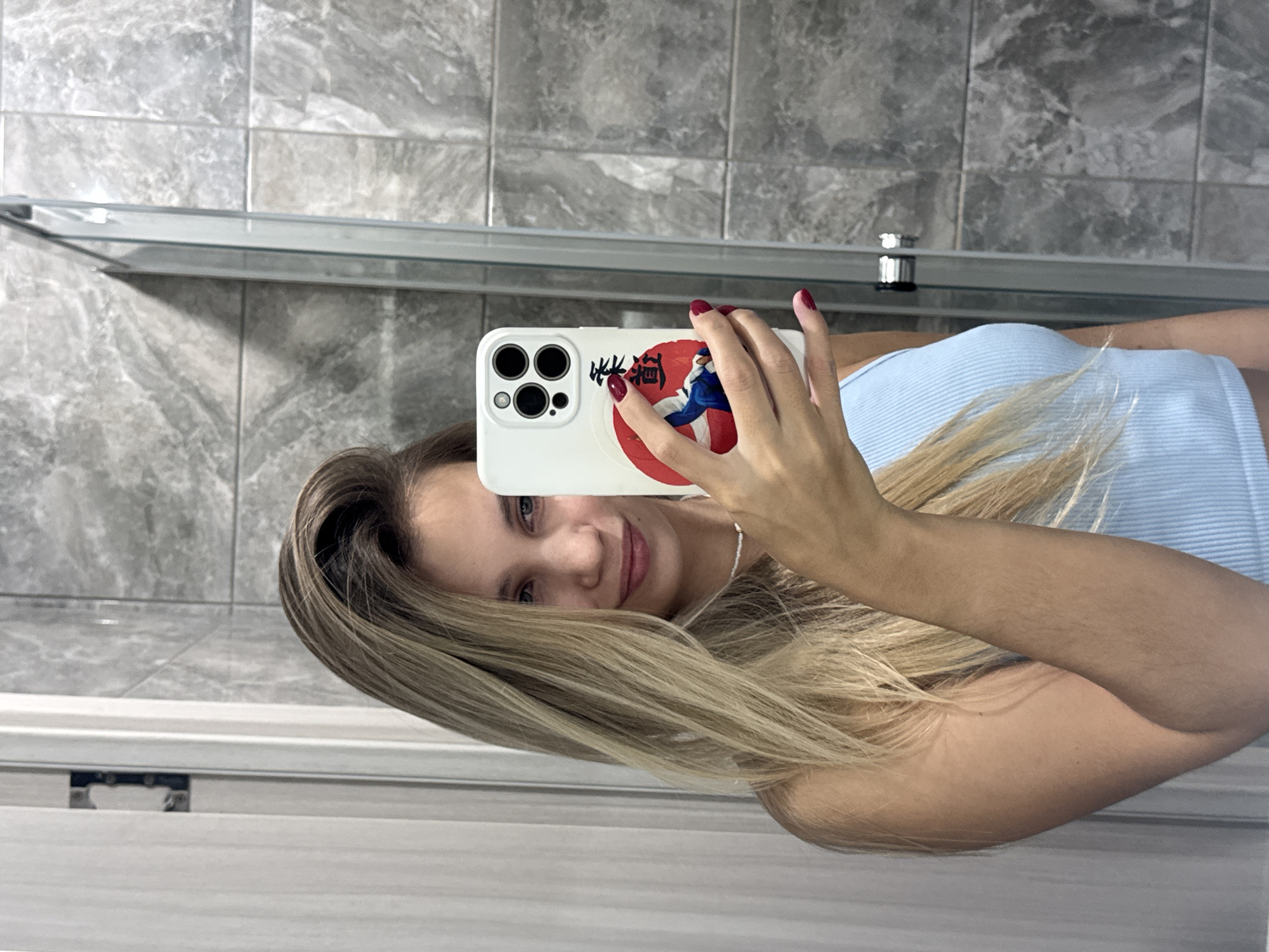 A young woman with long blonde hair, wearing a light blue ribbed tank top, stands in front of a bathroom mirror, taking a selfie. Her hair is partially covering her face. She is holding a white smartphone with a sticker on the back depicting a Japanese motif. Her fingernails are painted red. The bathroom features grey tiled walls and a glass shower enclosure. The scene is indoors, likely a residential bathroom. There are no indications of Chaikovsky, Russia, in the image.