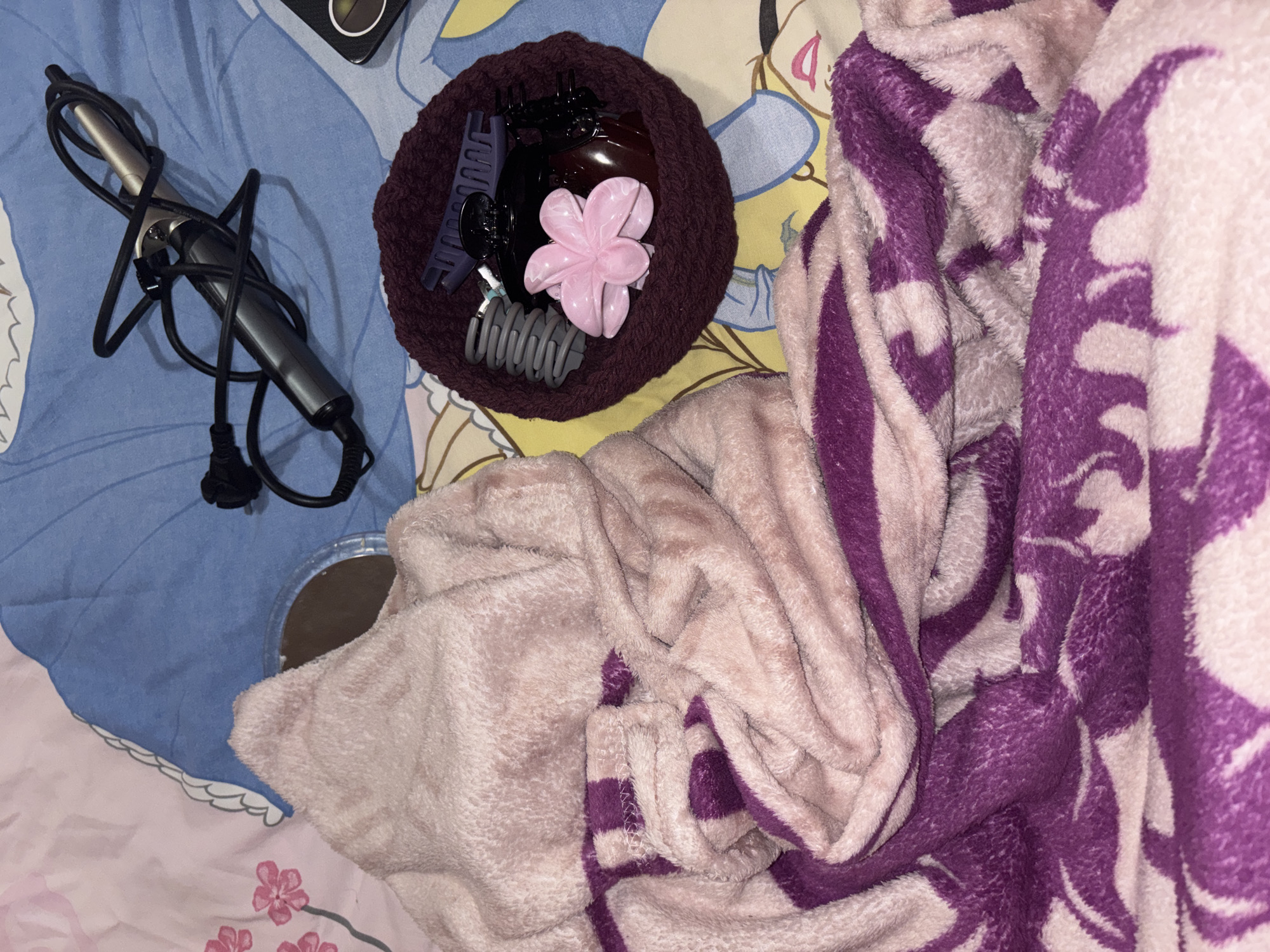 The image shows a bed in Garmen, Bulgaria, with a light blue and pink patterned bedsheet partially covered by a light pink and purple fuzzy blanket. A curling iron with a tangled black cord sits on the left side of the bedsheet. Next to it is a brown knitted bowl containing hair accessories, including claw clips and a flower-shaped clip. A small round mirror is positioned near the edge of the blanket. No people are visible in the image.