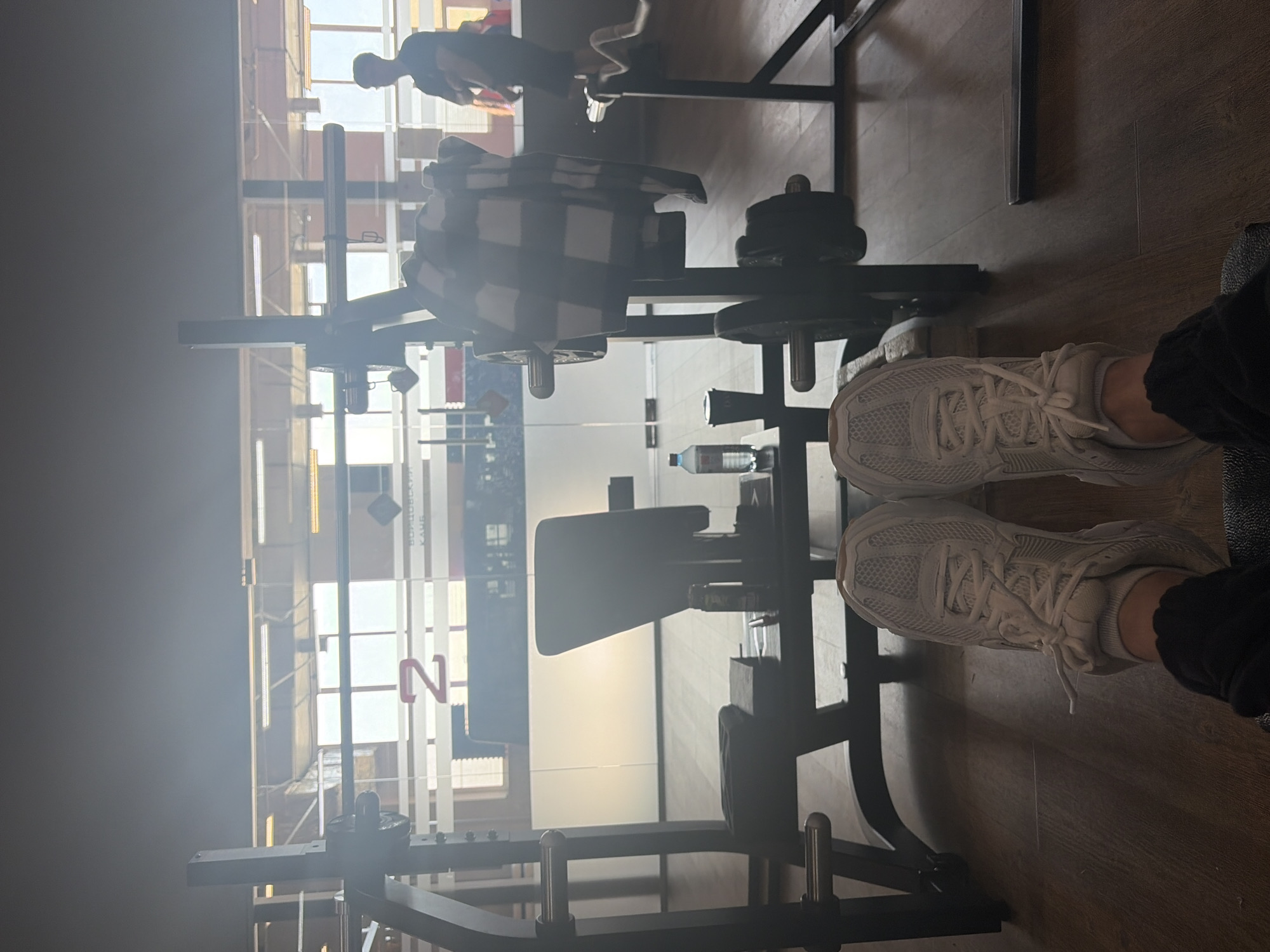 This image captures an indoor scene within a gym or fitness center, presented from a first-person perspective. The camera is positioned low, focusing on the photographer's feet in the foreground, which are clad in white athletic sneakers and dark pants, resting on a light brown wooden floor.
In the mid-ground, various gym equipment is visible, primarily a black weight rack or machine. Several black weight plates are stacked on the equipment, and a black and white plaid shirt or towel is draped over one section. A small water bottle sits on a lower part of the equipment.
The background is brightly lit, likely by large windows or glass partitions, causing significant glare and reflections that create a hazy effect across the upper portion of the frame. Through this light and haze, another person wearing dark clothing is faintly visible, standing or sitting further back in the gym. Vertical white panels are also present in the background, one of which displays Cyrillic text. The text reads "ВЕРОВОЙ КЛУБ" (VEROVOY KLUB), translating to "Faith Club" or possibly a localized name, with a prominent red "N" and potentially a "2" below it.
The strong natural light indicates it is daytime. The atmosphere is casual, suggesting a moment of rest or observation during a workout session. The setting, with its equipment and visible text, places the location in a fitness facility in Chaikovsky, Russia.
