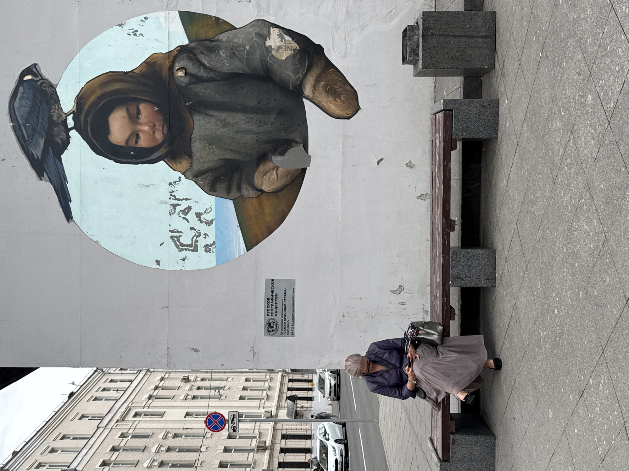 The image captures a street scene in Moscow, Russia. On the right side of the frame, a mural is painted on a white wall, depicting a child wearing a fur-lined hood and coat, with a bird perched on the child's head. The mural is set against a light blue sky with hints of water and a brown landscape. A poster, appearing to be an advertisement or announcement for a photo contest by the Russian Geographical Society, is affixed to the wall beneath the mural. In the foreground, an elderly woman with grey hair is seated on a wooden bench with granite supports, looking at her smartphone. She is wearing a dark purple jacket and a long, flowing grey skirt. Beside her on the bench is a metallic handbag. The bench is positioned on a paved sidewalk. To the left of the bench, a street with cars and a traffic sign is visible. The overall environment suggests an urban setting in daylight.