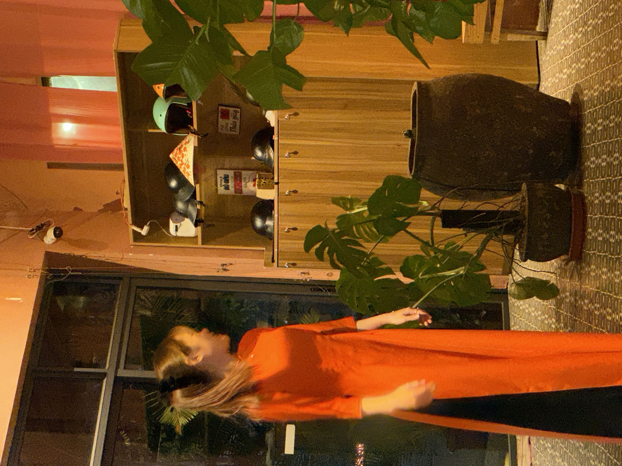 The image depicts an indoor scene with a young woman in an orange Áo dài standing by a large window. She has her hair pulled back in a ponytail and is looking to her left. Behind her, a wooden shelving unit displays several helmets, including a blue one, an orange one, and black ones. There is also a conical Vietnamese hat on the shelf. A CCTV camera is visible on the wall above the shelves. In the foreground, two potted plants, one with large leaves, are placed on a patterned floor. A large, dark clay pot sits to the right of the plants. The overall ambiance suggests a casual establishment, possibly a shop or a cafe, in Đà Nẵng, Vietnam.