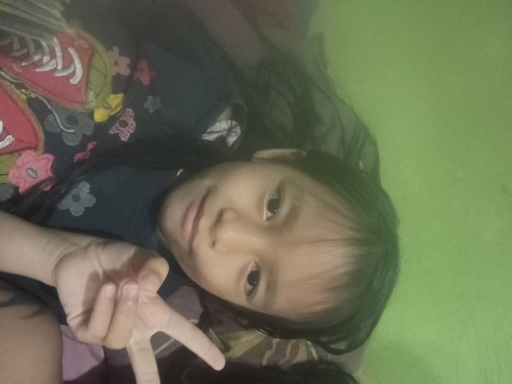 The media file is a selfie of a young girl lying down. She is wearing a dark blue shirt with a floral pattern. Her right hand is raised, making a peace sign. The background is a solid light green, suggesting an indoor setting like a bedroom. The lighting is dim, and there's a slight green cast over the image, indicating artificial lighting or a camera filter. The girl's face is the primary focus, and she appears to be looking at the camera with a neutral expression. The overall mood is casual and relaxed. The time of day and weather are not discernible from the image. There is no visible text in the media file. Given the provided location context of Pagar Alam, Indonesia, this could be a personal photograph taken in a home environment.