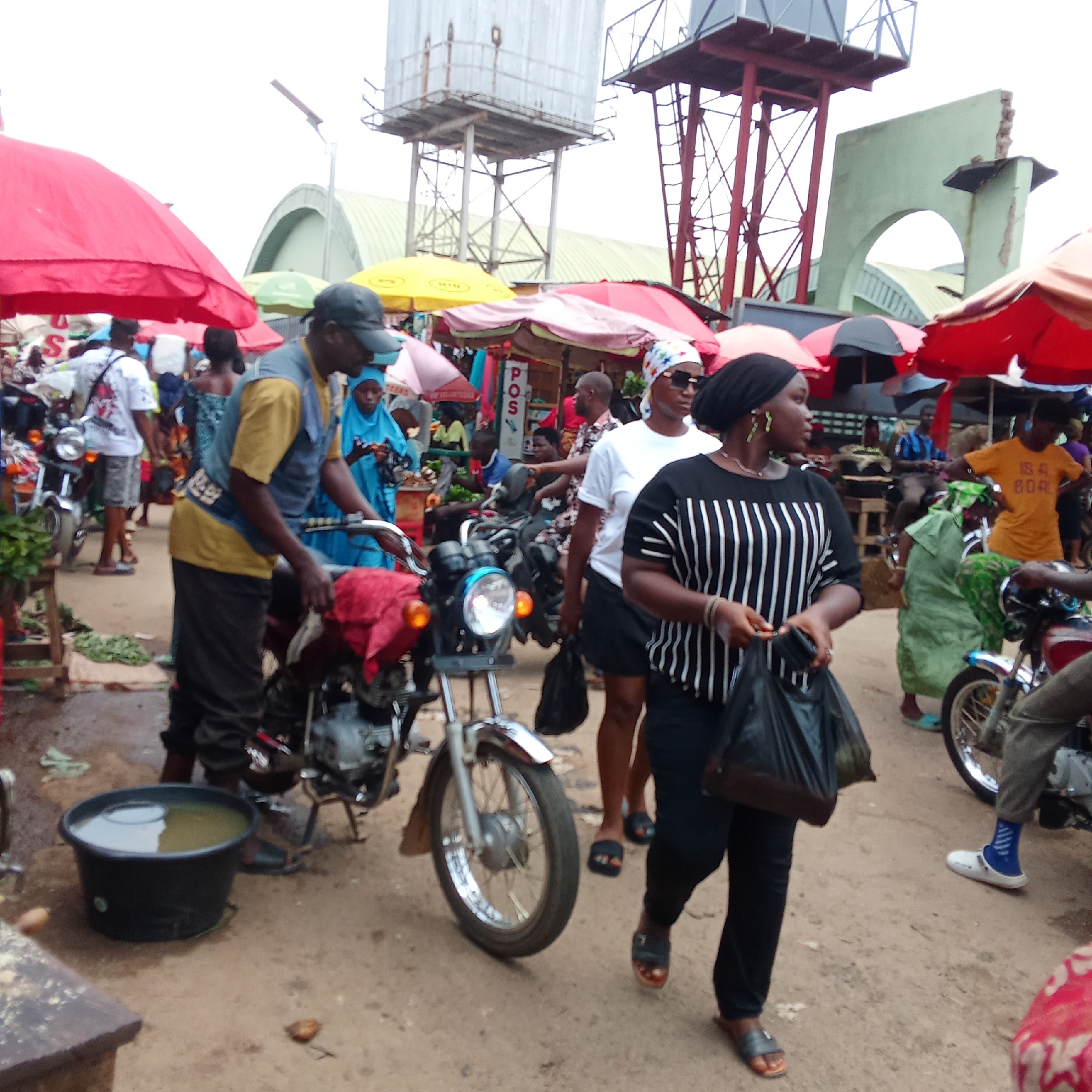 The image depicts a bustling scene in what appears to be a marketplace in Akwanga, Nigeria. Several people are present, including a woman in a black and white striped shirt walking with a black plastic bag, and a man in a yellow shirt attending to a motorcycle. Other individuals are visible in the background, engaged in various activities, suggesting a busy market day. There are also several umbrellas overhead, providing shade for vendors and shoppers. The environment includes a bucket of water near the motorcycle and a dirt ground. In the background, there are metal structures, including water towers and part of a building or gateway with a damaged arch. The overall scene indicates a vibrant commercial activity typical of a West African market.
