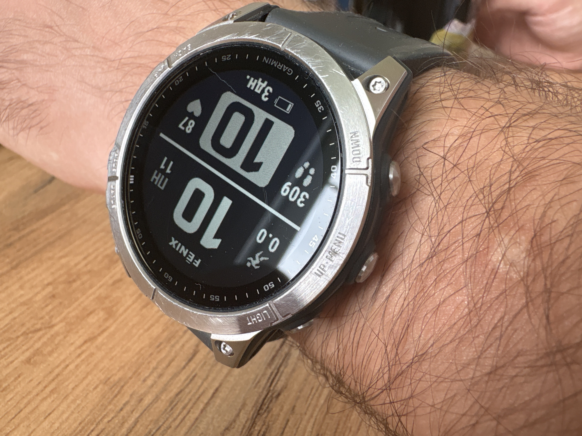 A close-up shot depicts a Garmin Fenix smartwatch worn on a hairy human arm. The watch face displays the time as 10:10, along with other data such as heart rate (87), steps (309), and distance (0.0). The watch has a silver-colored bezel with visible scratches and a black wristband. The buttons are labeled "LIGHT," "UP-MENU," and "DOWN." The background shows a wooden surface. There is no evidence to pinpoint the location as Kazan, Russia.