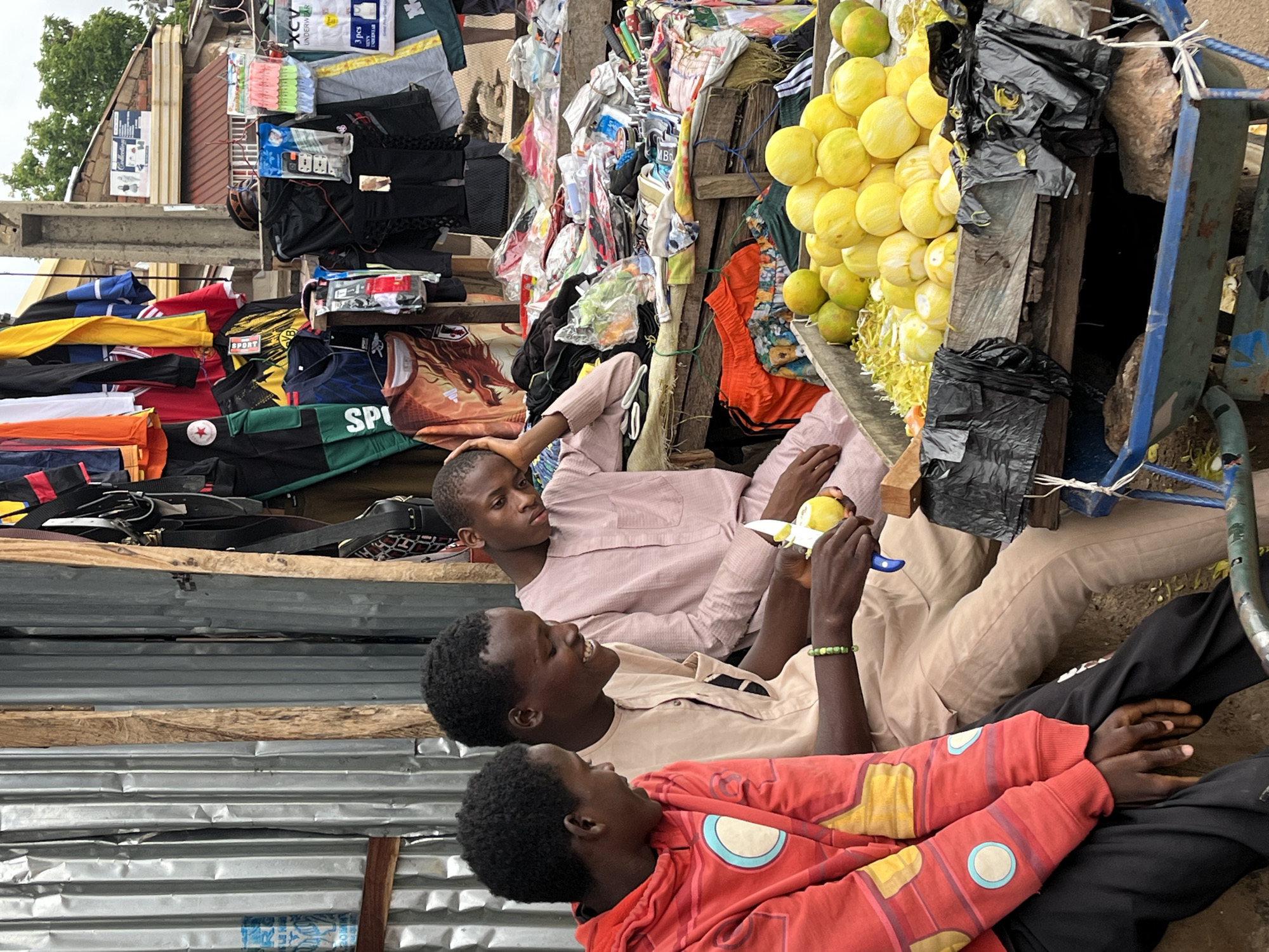 Three young men are seated in front of a market stall in Gura Top, Nigeria. The man on the left wears a red hoodie with yellow and blue circular accents. He is looking down and to his right, with his hands resting on his lap. The man in the middle is wearing a light brown, long-sleeved tunic and is smiling, looking to his left. He is peeling a yellow fruit with a small knife, and a green beaded bracelet is visible on his left wrist. The man on the right, also in a light brown tunic, is leaning his head on his hand and looking downwards, appearing pensive.
The market stall behind them is laden with various items, including clothing hanging on display and a large wooden crate filled with yellow fruits, possibly lemons or oranges, and some green fruits. The stall itself is constructed from corrugated metal and wood. In the background, a building with a red roof is visible. The ground appears to be dirt.