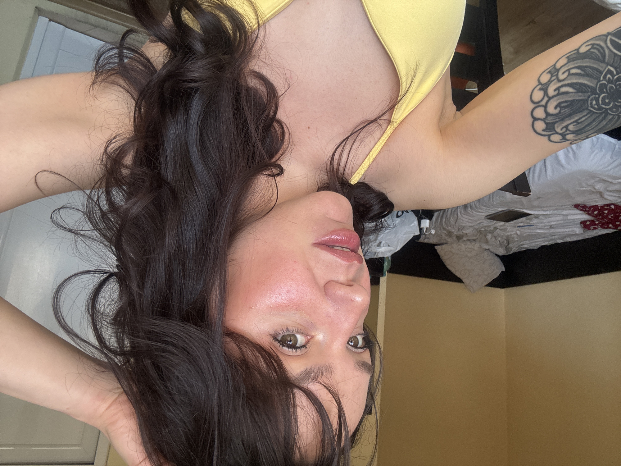 A woman with dark brown hair and fair skin is positioned at an angle, facing the camera. She is wearing a yellow top, and a tattoo is visible on her upper left arm. The background shows a beige wall in what appears to be an interior setting. There is no evidence to place this image in Blagoevgrad, Bulgaria.
