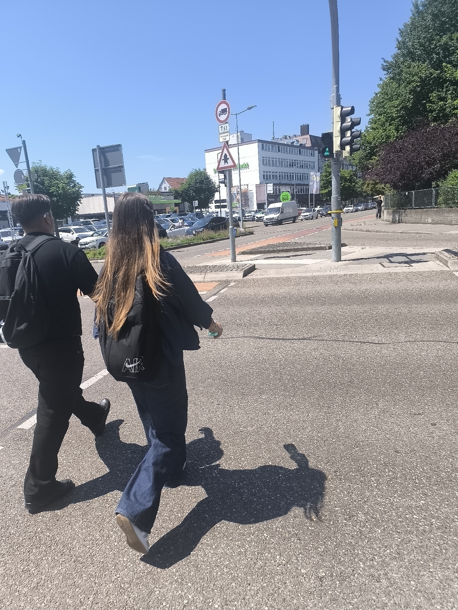 Two individuals are walking across a crosswalk in an urban environment. The person on the left is wearing black clothing and a backpack, while the person on the right is wearing dark blue jeans, a dark jacket with an "AIR" logo, and also has a backpack. The scene appears to be a sunny day with a clear blue sky. In the background, there are buildings, trees, traffic lights, street signs, and parked cars. The crosswalk is paved with a rough, speckled material.