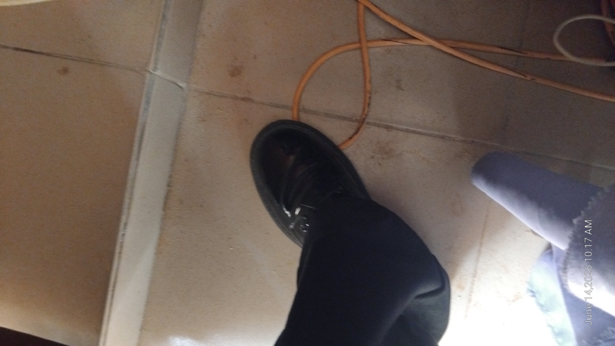A person's left foot, wearing a black, shiny shoe and dark pants, is positioned on a tiled floor. An orange electrical cord is looped on the floor near the shoe. In the lower left corner, a blue, rolled-up item is partially visible. The timestamp "June 14, 2026 10:17 AM" is present in the bottom left of the image. The environment appears to be indoors, on a floor with visible grout lines and some dirt or scuff marks. No specific landmarks indicating the city of Jalingo, Nigeria, are discernible from the image.