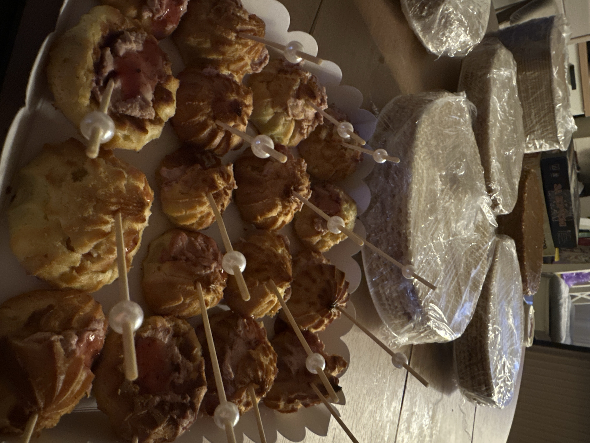 A close-up, high-angle shot shows a table laden with baked goods. In the foreground, a white, scalloped-edge tray holds numerous small, round pastries filled with a pinkish cream, each pierced with a wooden skewer topped with a pearl bead. Behind this tray, several large, round cake layers are stacked and wrapped in clear plastic. To the left and right of these cake layers, more plastic-wrapped cake layers are visible. Further back, a light brown cake with a smooth surface sits on the table. The setting appears to be indoors, with a wooden table surface and a hint of furniture in the background. The image captures a preparation scene for a bakery event or celebration.