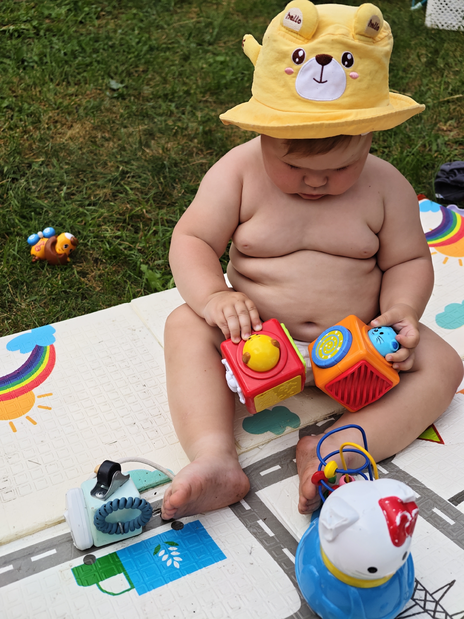A baby wearing a yellow sun hat with bear ears and the word "hello" on it sits on a play mat outdoors. The baby is naked from the waist up and is holding two colorful toys, a red square with a yellow sphere and a blue and orange cube with a blue animal head. To the left of the baby is a brown toy bug with blue wings. Behind the baby is a grassy area. The play mat is decorated with a rainbow, a sun, a road, and a blue square with white leaves.