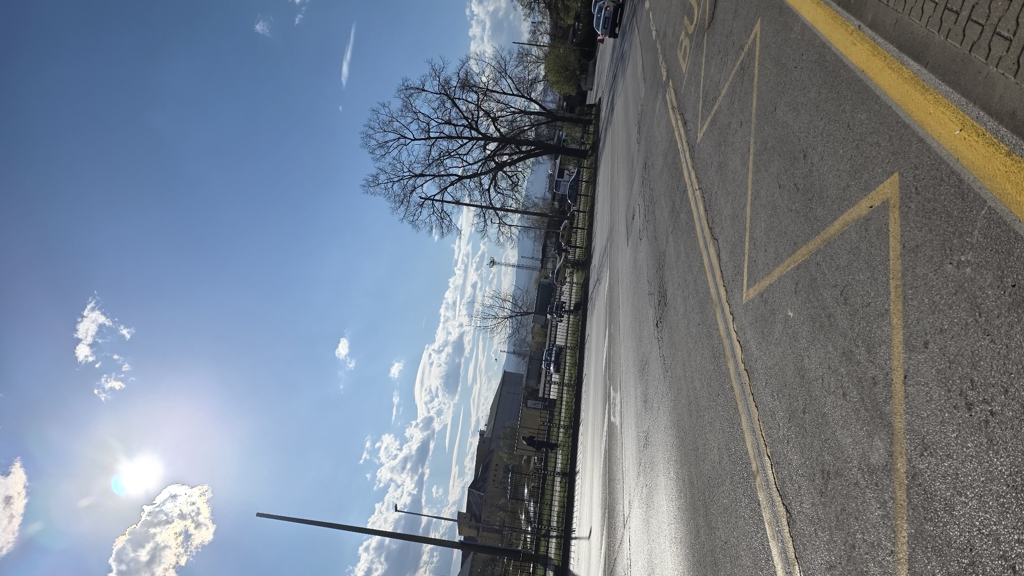 The image presents a street view on a sunny day in what appears to be an urban area. A road with yellow painted lines and markings occupies the right side of the frame, suggesting it is a bus stop zone. The road surface is asphalt and slightly wet. The left side of the frame features a bright blue sky with scattered white clouds and the sun shining brightly. Buildings, bare trees, and a metal fence are visible along the horizon line. Cars are parked along the side of the road further in the background. Given the mention of Blagoevgrad, Bulgaria, it can be inferred that this is the location.