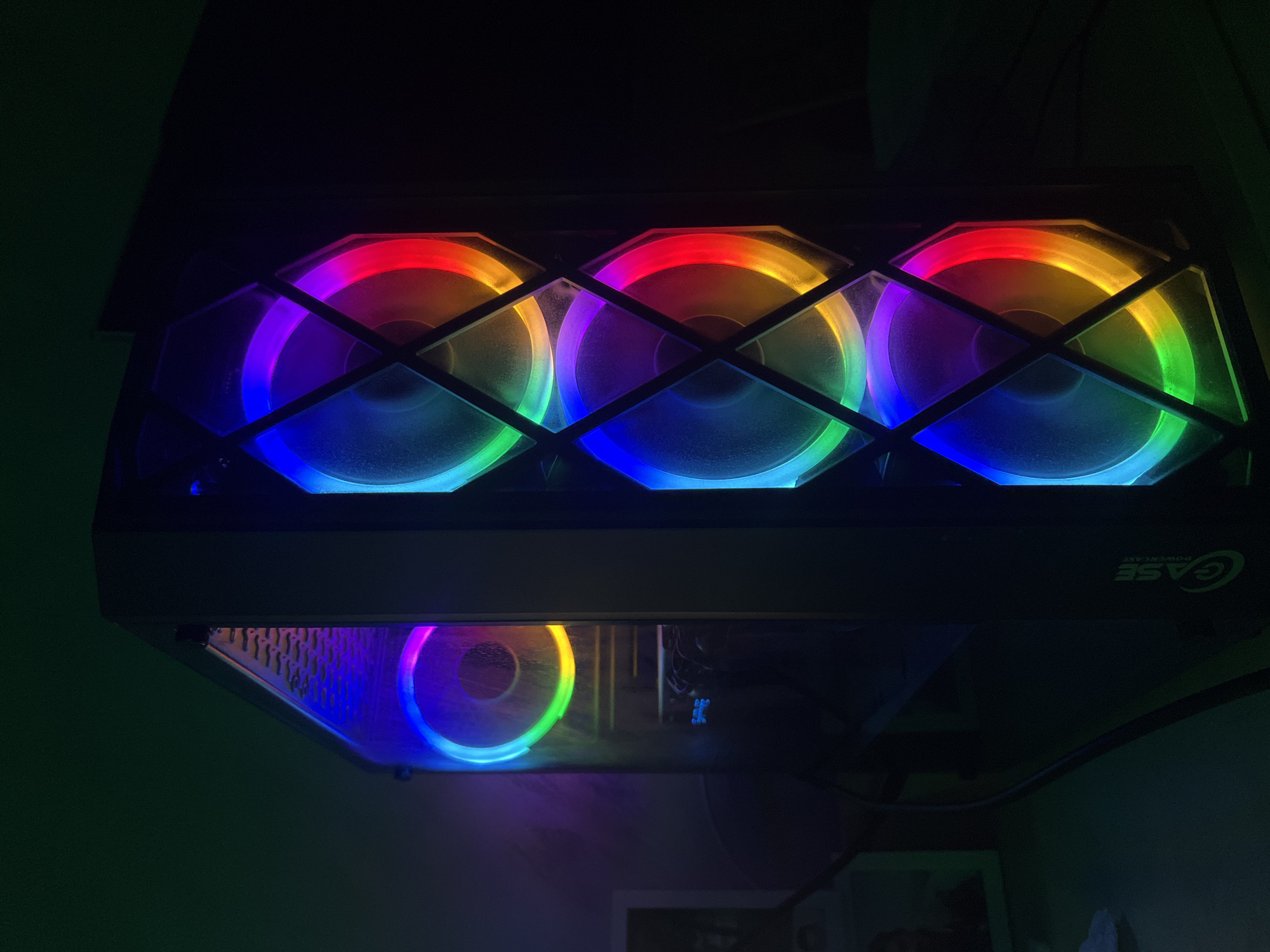 The image shows a black computer tower with illuminated RGB fans. The computer is placed on a desk in a dimly lit room, with a green wall in the background. The computer tower has a glass side panel, through which a fan with a colorful RGB ring is visible. The front of the tower features three more fans, also with RGB lighting. The lighting is a gradient of colors, including blue, purple, red, and yellow. A cable is visible running from the computer to the right. There is a faint reflection of a person on the glass panel. The logo "CASE POWERPLAY" is visible on the lower right side of the computer tower. There is no indication of the location being Blagoevgrad, Bulgaria.