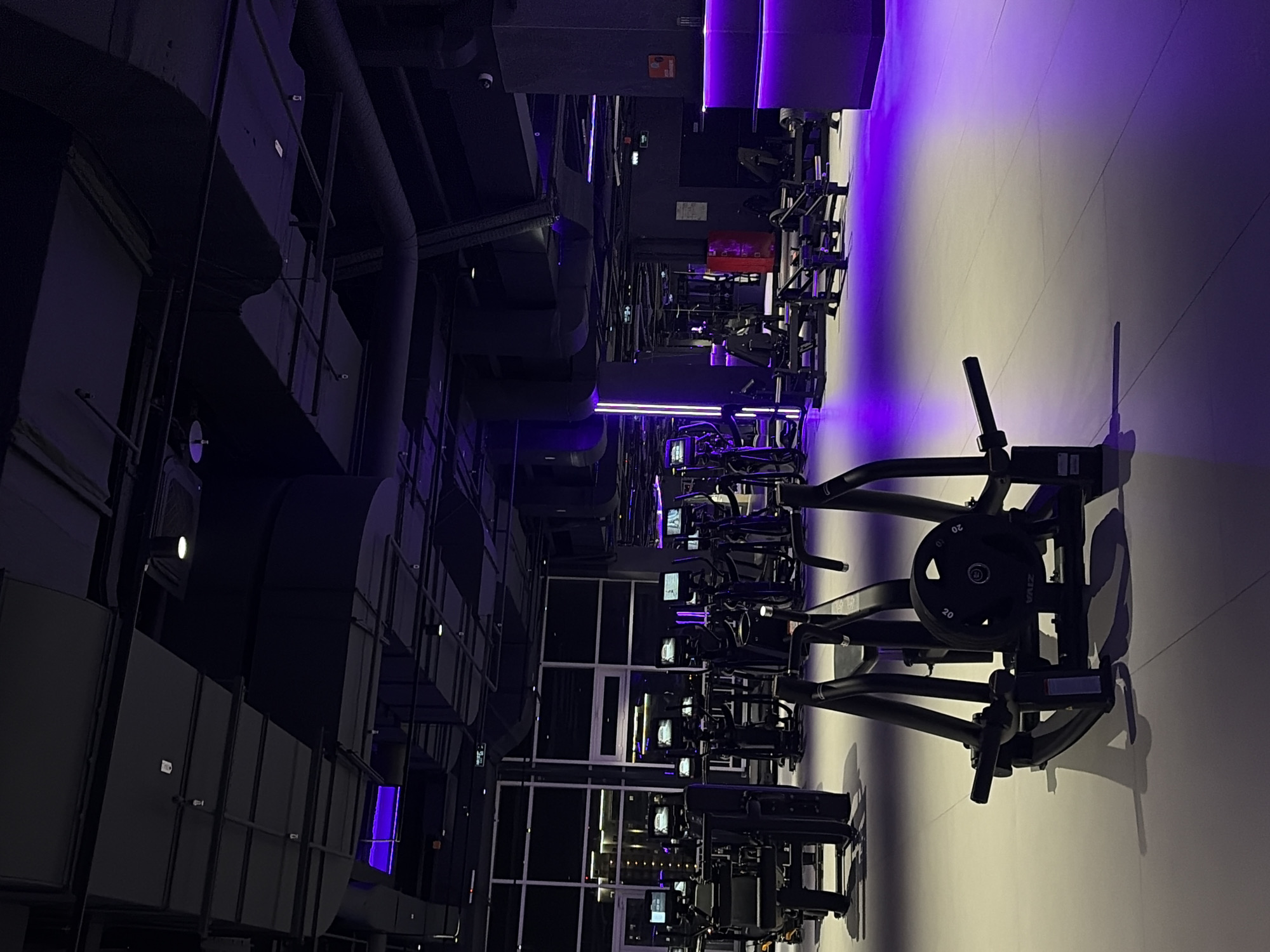 The image shows the interior of a modern, well-lit gym. The space is primarily characterized by its extensive collection of exercise equipment, including treadmills, ellipticals, and weight machines, all rendered in dark, sleek materials. The lighting scheme is predominantly purple, casting a vibrant and atmospheric glow across the entire space, particularly on the floor which appears to be a light-colored, smooth material. The ceiling features industrial-style ductwork and lighting fixtures, contributing to a contemporary aesthetic. Large windows line one side of the gym, offering a glimpse of an urban cityscape at night, with illuminated buildings visible in the background. The overall impression is that of a high-end fitness facility designed for intense workouts, with the purple lighting suggesting a stylish and energetic ambiance. The image was taken in the city of Krasnoyarsk, Russia.