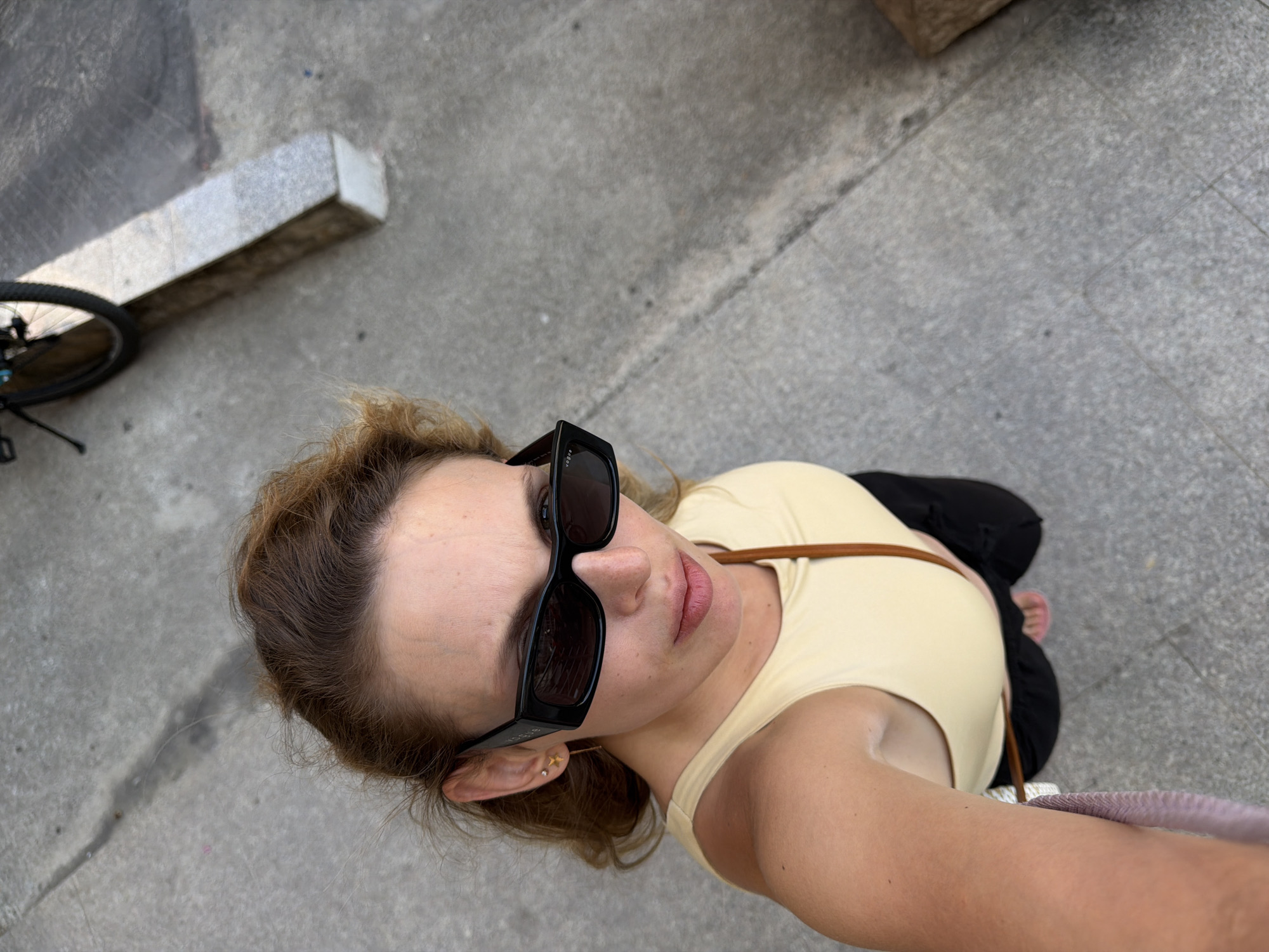 The media file shows a woman taking a selfie outdoors, likely in Budva, Montenegro. She is wearing sunglasses and a light yellow top, with black pants visible. Her hair is light brown and appears slightly windblown. She is wearing a small star-shaped earring. A brown strap from her bag is visible across her chest. The background is a concrete sidewalk with some visible cracks. To the left of the frame, a bicycle wheel and part of the frame are visible, suggesting the woman may have arrived by bicycle. A stone block or curb is also present in the background. The lighting suggests it is daytime. The woman's expression is neutral, and the overall mood of the image is casual and candid.
