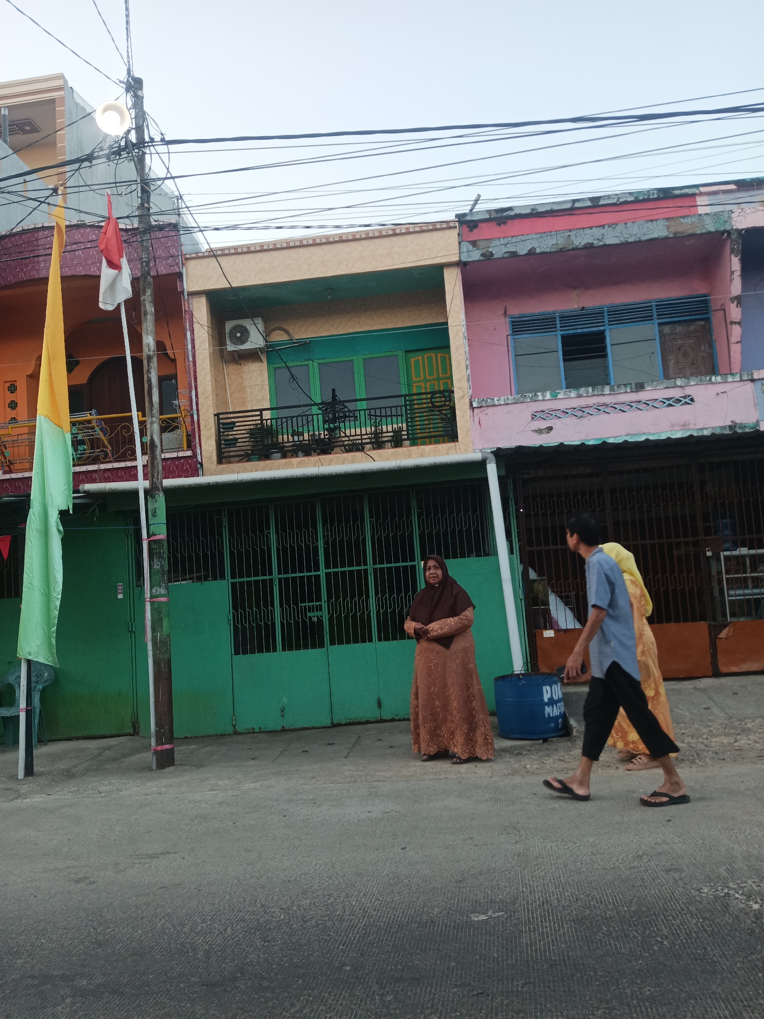 The image depicts a street scene in Pangkajene, Indonesia, with several colorful buildings lining the right side of the frame. In the foreground, a paved road runs horizontally. On the left side of the road, a flag pole with red and white Indonesian flags and a yellow and green flag is visible. Behind the flagpole are green doors and an orange building. To the right of the flagpole, a woman in a brown hijab and a patterned dress stands near a blue barrel. Further to the right, two individuals, one in a blue shirt and black pants and the other in a yellow outfit, are walking away from the camera. The buildings behind them are multi-story structures with various colored facades, including orange, tan, and pink. Electrical wires and a street light are prominent above the buildings. The sky is overcast.