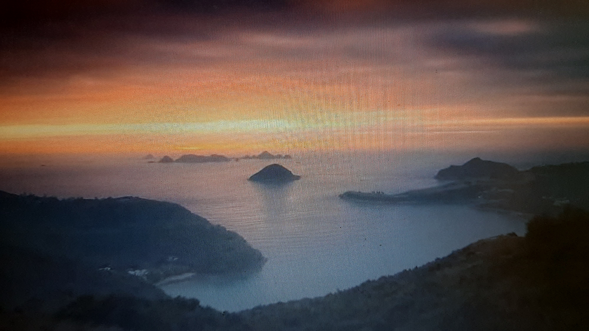 The image depicts a dramatic sunset over a coastal landscape with several islands scattered in the calm sea. The sky is ablaze with vibrant hues of orange, yellow, and purple, casting a warm glow on the water and surrounding land. Dark, silhouetted mountains rise from the sea, their peaks partially obscured by mist. In the foreground, the edge of a dark, forested mountain slopes down towards the water, with glimpses of what appear to be small structures or lights nestled within the vegetation. The overall scene evokes a sense of serene beauty and remoteness.