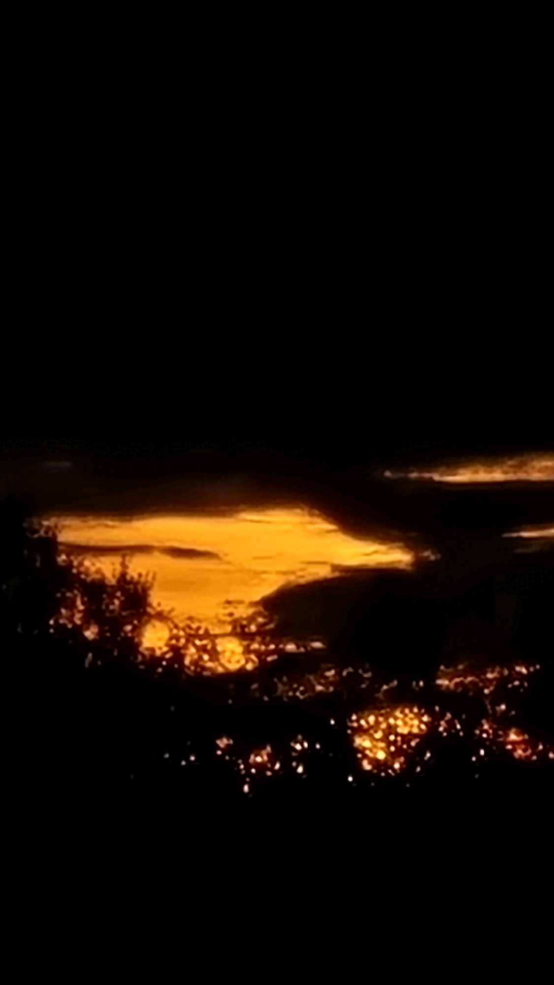 The image depicts a twilight scene over the city of Vysokoe, Russia. The sky is dominated by dark clouds, with a band of bright orange light illuminating the horizon, suggesting the last rays of sunset. Below the sky, a silhouette of trees and vegetation is visible in the foreground on the left. Further into the distance, the lights of the city are scattered across the landscape, appearing as small, glowing orbs against the darkening terrain. The overall impression is one of a city transitioning from day to night, with the natural beauty of the sky contrasting with the man-made illumination below.