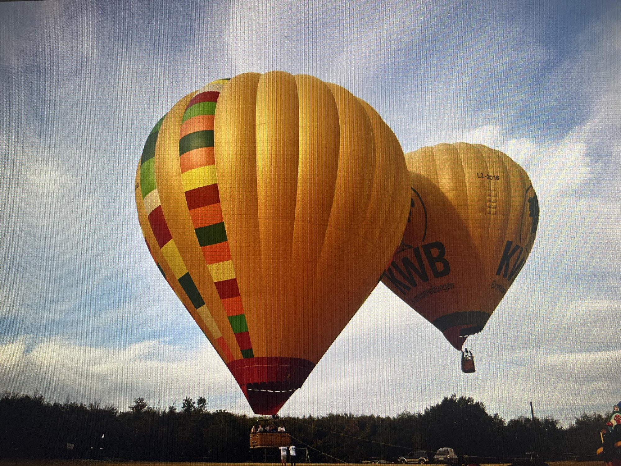The image captures an outdoor hot air ballooning event, likely during preparation or landing, set against a partly cloudy sky in Varna, Bulgaria.
The primary subjects are two large hot air balloons. The one in the foreground is predominantly yellow/gold, featuring vibrant vertical stripes of red, green, orange, and lighter yellow. Its basket is visible at the bottom, holding several people, while two individuals stand on the ground nearby, possibly handling ropes. This balloon appears to be tethered or very close to the ground.
Behind and slightly to the right of the first balloon is a second, mostly plain yellow/gold hot air balloon. On its surface, the text "KWB" and "Biomasseheizungen" is clearly visible, suggesting sponsorship. Further up, the registration number "LZ-2016" can be seen. This balloon's basket is also visible, suspended below it, and it appears to be either ascending or descending, or already airborne at a lower altitude than typical flight.
The setting is an open field or clearing, with a dark green treeline forming the horizon. The sky above is a mix of blue and white, with scattered clouds. The bright, natural lighting indicates it is daytime, likely late morning or afternoon. The overall scene suggests a lively and engaging recreational event. People's relaxed postures indicate a peaceful atmosphere, typical of such gatherings.