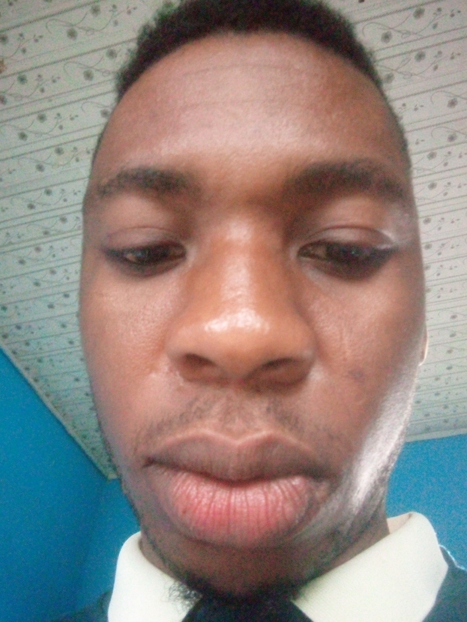 The image is a close-up selfie of a Black man wearing a white collared shirt. He is looking down, and the camera angle is tilted upwards. The background includes a patterned ceiling and a blue wall. There is no evidence in the image to suggest the location is in Ikorodu, Nigeria.
