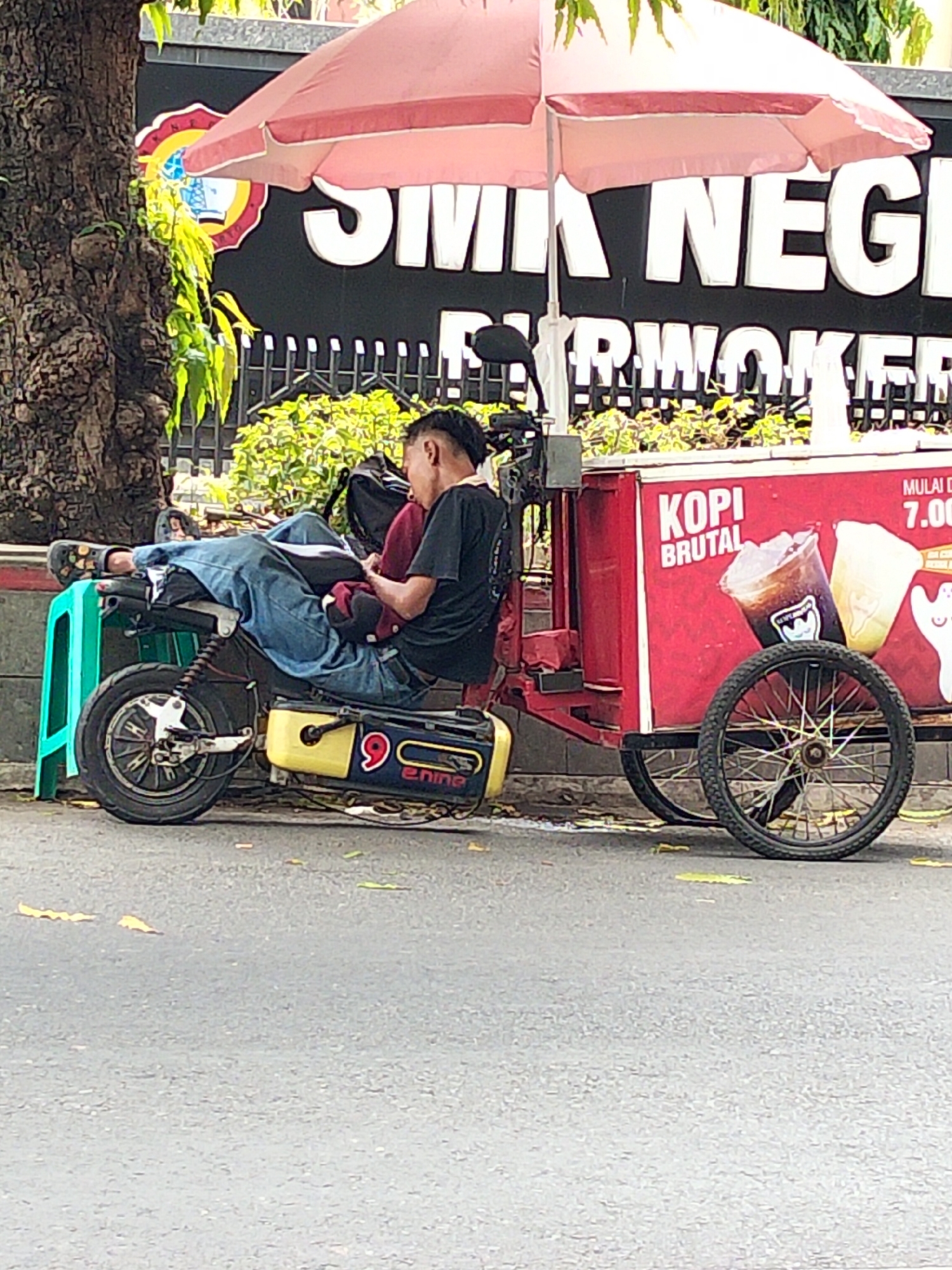 The image depicts a street scene in front of a building with large white lettering on a black background, partially obscured by a pink umbrella. The lettering appears to read "SMK NEGERI" followed by more text, possibly indicating a school. To the left of the building is a large tree trunk. In the foreground, a person is resting on a red cart with a black umbrella overhead. The cart has a graphic of two drinks and text that says "KOPI BRUTAL" and "MULAI 7.00". The cart is attached to a wheeled mechanism. The person is wearing a black t-shirt and blue jeans, with their legs propped up on a green stool. A dark backpack is behind them. To the right of the cart is a large wheel. The ground is asphalt, with some fallen leaves. The overall scene suggests an outdoor vendor stall or resting area.
