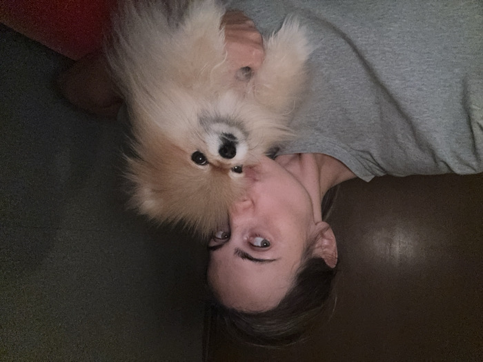 An indoor photograph captures an adult female holding a small, light brown Pomeranian-type dog. The female, positioned on the left, wears a grey crew-neck t-shirt. She is angled to her right, with her head tilted and lips pursed towards the dog's head in a kissing gesture. Her visible right eye glances towards the left side of the frame. Her hair, light brown, is pulled back. The dog, held in her arms, faces forward with a neutral expression. Its fur is fluffy and predominantly light brown with lighter highlights. The background consists of a dark, undifferentiated wall, possibly dark grey or brown, indicative of an interior space with subdued lighting. A small portion of a red surface, likely furniture, is visible in the lower right corner. No specific visual indicators within the photograph identify a precise address or landmark within the city of Warsaw, Poland.