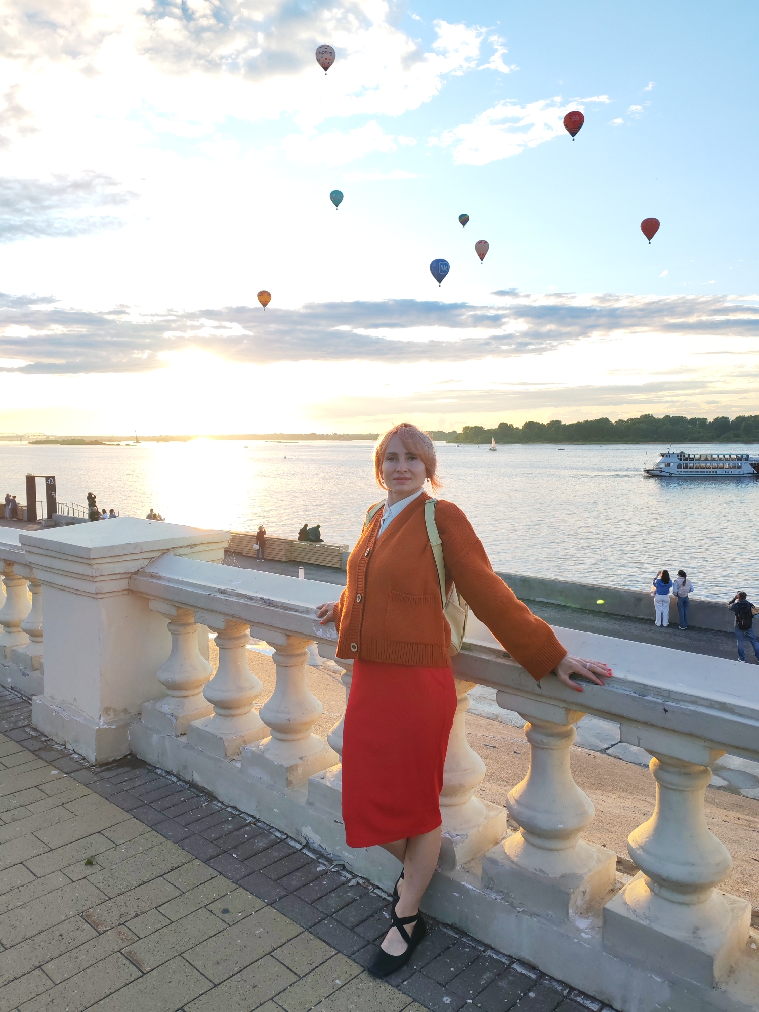 A woman with pink hair and an orange cardigan over a white shirt and red skirt poses for a photo on a promenade next to a decorative balustrade. The sun is setting over a wide river, casting a golden glow on the water and sky, which are dotted with several colorful hot air balloons. In the background, a large white boat with multiple decks sails on the river, and the opposite bank is lined with trees. Several other people are visible on the promenade, some watching the balloons, others looking at the river. The overall mood is peaceful and festive, likely during a hot air balloon festival. The time of day is sunset.