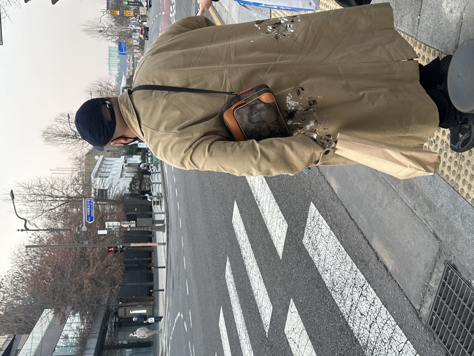 A person is depicted from behind, standing on a sidewalk curb at a street intersection on Itaewon-ro 55-gil in Seoul, South Korea. The individual wears a dark blue baseball cap, a black face mask, a long tan trench coat, black pants, and black athletic shoes. The lower right side of the trench coat exhibits multiple white and black paint splatters. A brown and black patterned crossbody bag with a black strap is slung over the person's right shoulder, and a light brown paper bag is held in the left hand.
The person stands on yellow circular tactile paving at the edge of the sidewalk, facing a multi-lane asphalt road with a marked white pedestrian crosswalk featuring four cracked stripes. A metal grate is visible on the sidewalk in the foreground. A red traffic light is illuminated on the left side of the intersection. In the background to the left, a modern building with a dark-framed glass facade is visible, along with two other pedestrians walking on the sidewalk. To the right, a white multi-story building and bare deciduous trees line the street. Distant vehicle traffic is visible on the road under an overcast sky.