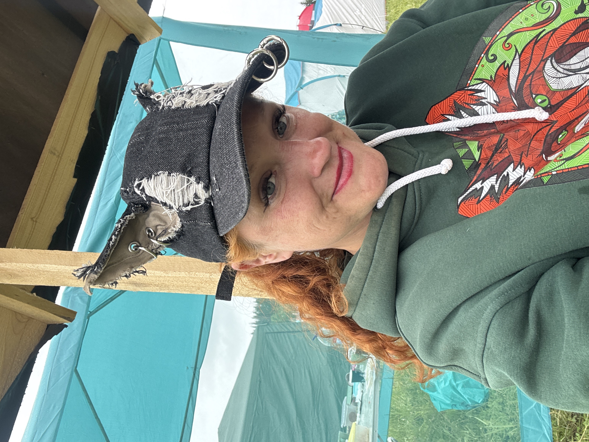 This medium shot, rotated 90 degrees clockwise, captures a smiling woman outdoors in Bol'shaya Kivara, Russia. She has vibrant, curly red hair and blue eyes, accentuated by dark eyeliner and mascara. Her lips are painted with red lipstick. She wears a dark green hooded sweatshirt featuring a prominent, stylized graphic of an animal head, possibly a fox or wolf, with green eyes and a detailed red and green pattern. A white drawstring hangs from the hoodie's neck.
On her head, she wears a distressed, dark denim baseball cap with frayed edges and two silver rings attached to the brim. Behind her, a wooden beam suggests she is under a temporary shelter or structure. The background reveals an outdoor setting with a large teal tent or canopy dominating the mid-ground, and other tents and green grass further back, indicating a camping trip or outdoor event. The diffuse lighting suggests it is daytime, possibly under an overcast sky. The woman displays a relaxed and friendly expression, looking directly at the camera. There is no visible text in the image.