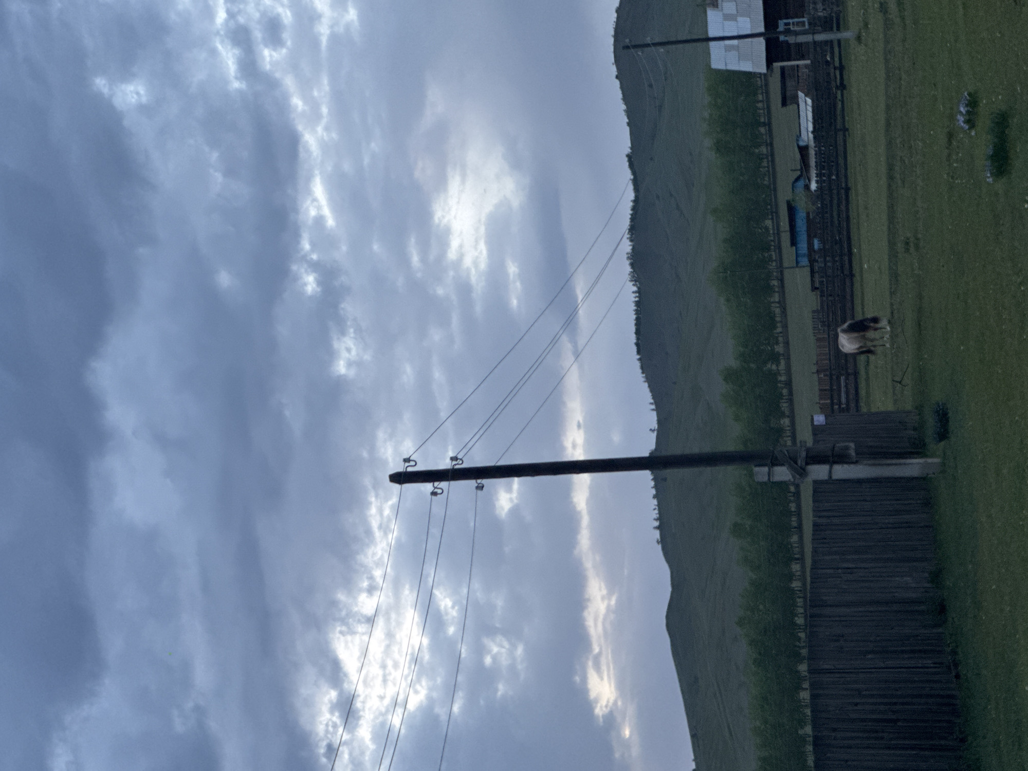 The image depicts a rural scene in Нижний Бургалтай, Russia. A large, green hill slopes upward in the background. In the foreground is a wooden fence running parallel to the base of the hill, with a utility pole and power lines extending into the cloudy sky. A single animal, possibly a horse or cow, stands in a grassy field near the fence. A few buildings are visible behind the fence. The sky is overcast with heavily clouded conditions.