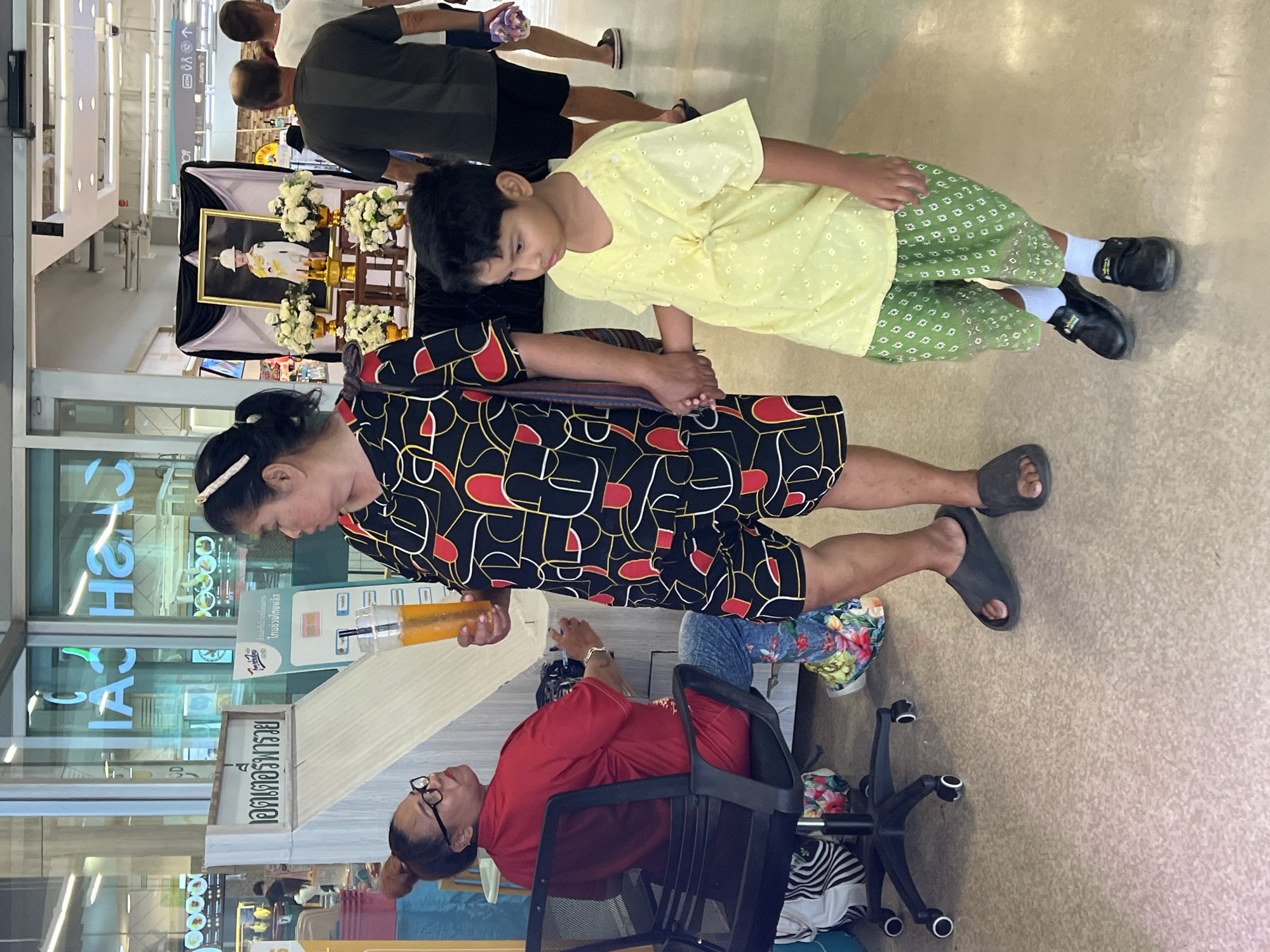 This image captures a scene inside a modern retail establishment, possibly a department store or supermarket, in Baan Lamai, Thailand. In the foreground, a woman in a black and red patterned dress stands holding a plastic cup with an orange beverage. She is accompanied by a young boy wearing a light yellow tunic and green patterned pants. They appear to be walking together, with the woman holding the boy's hand.
To the left, a woman wearing glasses and a red shirt is seated in a black office chair, looking towards the woman with the drink. She has a colorful, floral-patterned scarf or fabric draped over her lap. Behind her, part of a service counter or information desk is visible, featuring Thai text and images.
In the background, there is a framed portrait of a young person in a ceremonial outfit, displayed on a stand adorned with white flowers. This suggests a memorial or cultural event. Other shoppers are visible in the middle and far background, browsing or walking through the store. The environment is well-lit with fluorescent ceiling lights and has a polished floor. The overall impression is of a busy public space within a commercial setting.