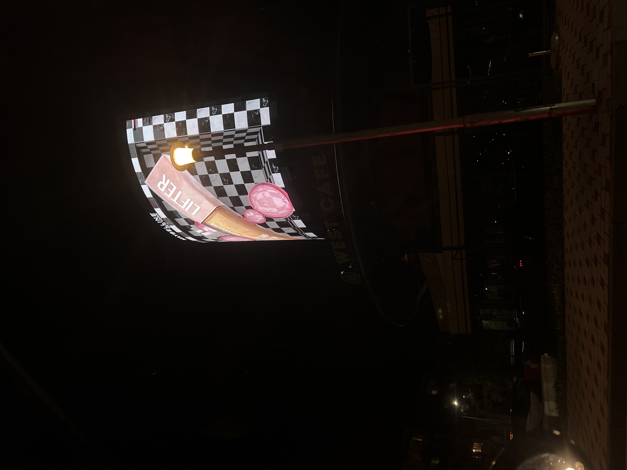 The image is taken at night. It shows a lit advertising sign for Maybelline Lifter Gloss outside West Cafe in Plovdiv, Bulgaria. The sign is checkered black and white and features an image of the product and pink lip shapes. The cafe sign is partially visible behind the advertising sign. The background is dark, with faint lights from the street. Part of a building's exterior wall is visible on the right.