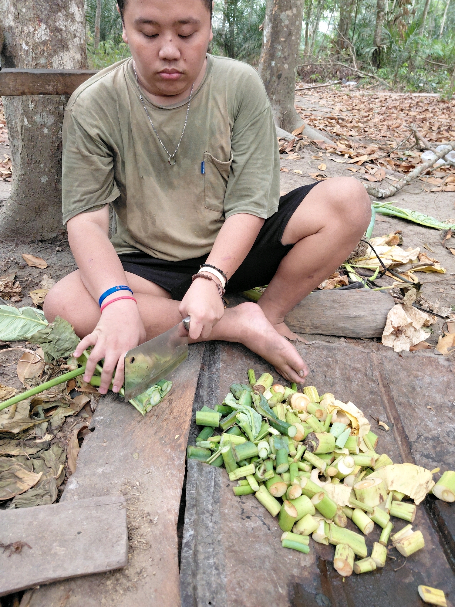 A young person with closed eyes sits cross-legged outdoors, in what appears to be a rural or forested area, possibly within Persiapan Mulya Raya, Indonesia. The individual is wearing a green t-shirt and black shorts, with colorful bracelets on their wrists. They are actively engaged in chopping green plant stalks with a large cleaver on a weathered wooden surface. A pile of chopped plant material has accumulated to the right of the person. The ground is covered with dry leaves, and the background shows trees and vegetation, suggesting a natural environment.