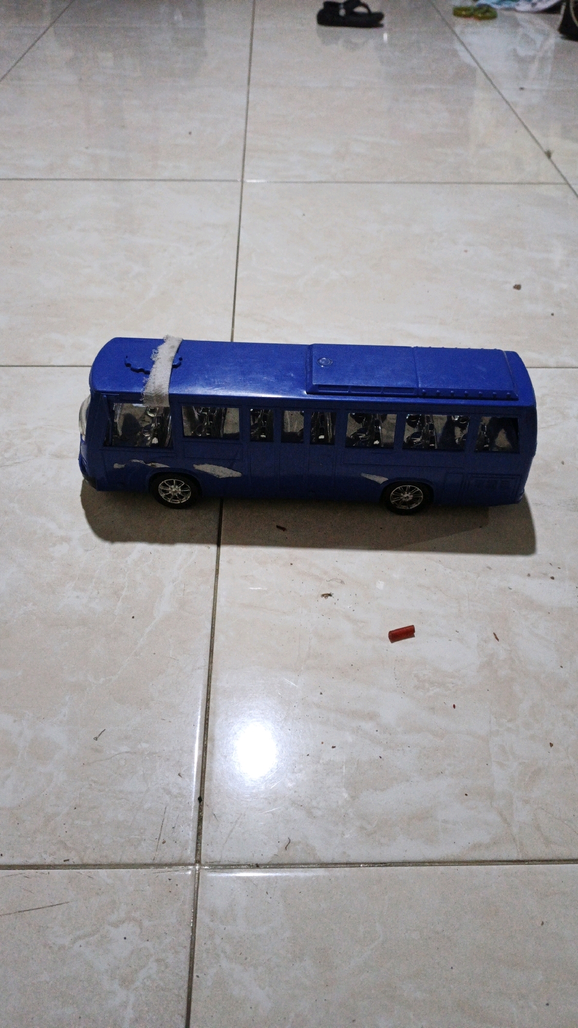 A blue toy bus is positioned on a tiled floor. The bus has visible windows and wheels. A white piece of tape is attached to the top of the bus. A small red object is on the floor near the bus. The floor tiles are light-colored with visible grout lines. The environment appears to be indoors, possibly a home or similar interior space, with the light suggesting artificial lighting. The location is not specified in Bono, Indonesia.