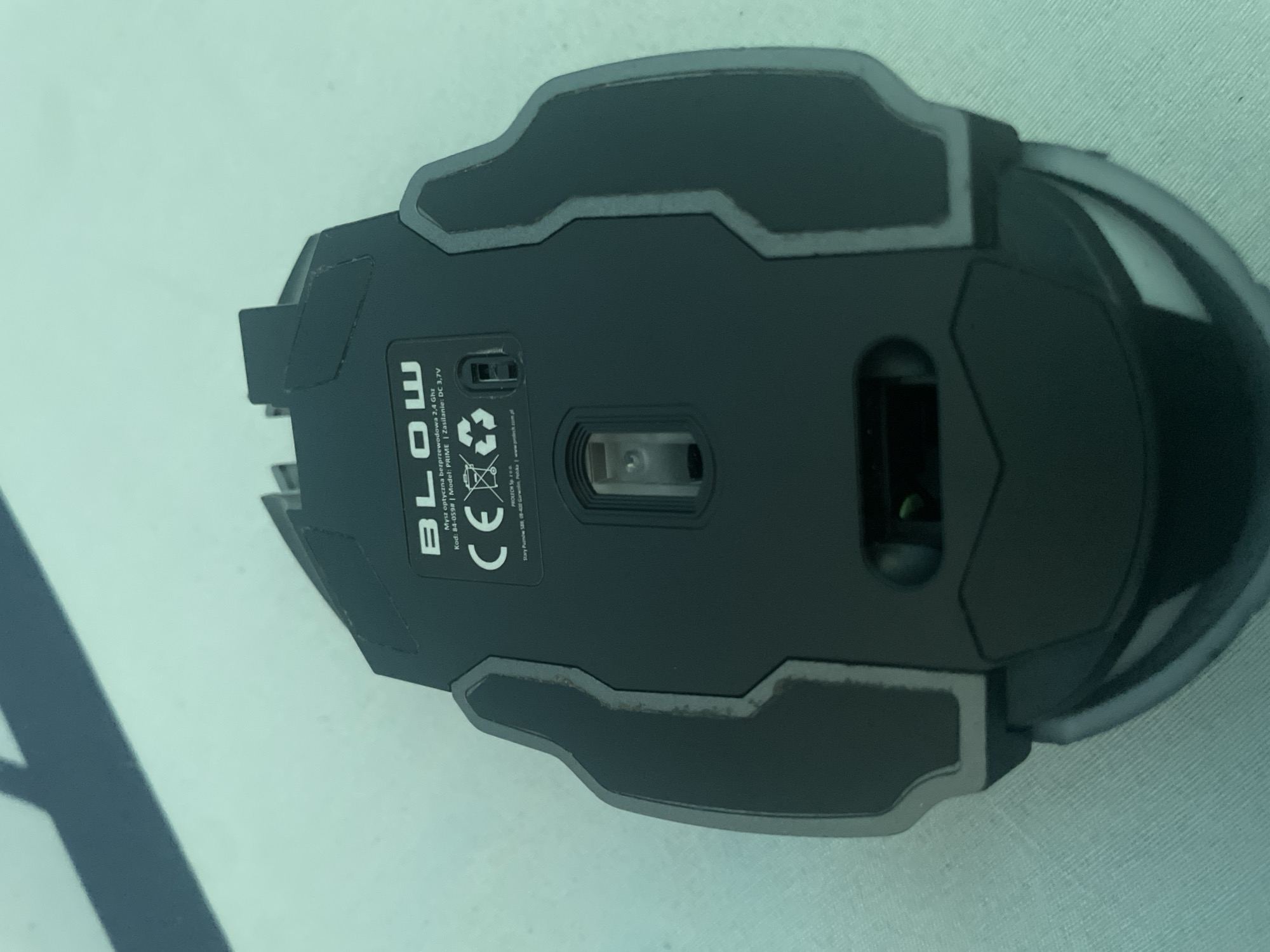 The image shows the underside of a black wireless computer mouse, likely manufactured by BLOW. The mouse is placed on a light-colored, textured surface, possibly a mousepad or fabric. Visible text on the mouse identifies it as a "Mysz optyczna bezprzewodowa 2,4 Ghz" (wireless optical mouse 2.4 GHz), with a product code "84-059#" and model name "PRIME". It also states the power requirement as "DC 3,7V". The CE marking is visible, indicating compliance with European standards. Below that, there is text that appears to be a company name and address in Kołobrzeg, Poland. The lighting suggests an indoor setting, possibly during the day. No people or specific events are depicted. The image focuses solely on the product.