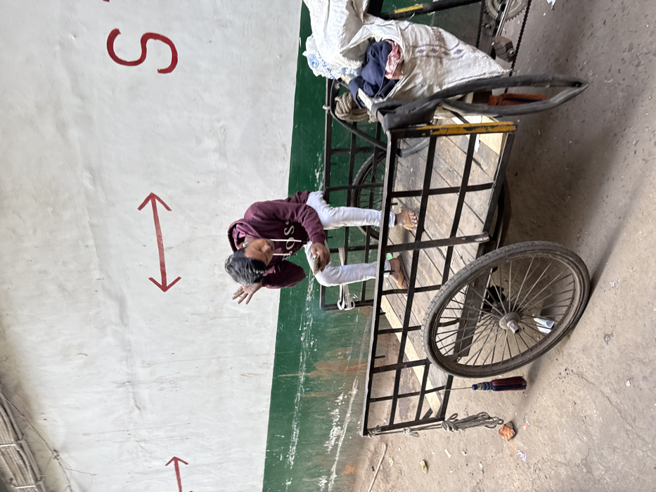 A young adult male is seated on the wooden flatbed of a black metal-framed cargo bicycle/rickshaw in an alleyway of Kolkata, India. He wears a maroon hooded sweatshirt displaying "BOSS," white pants, and brown sandals. His left hand holds a light-colored mobile device, while his right hand is raised towards his head with fingers splayed. The cargo vehicle's front left spoked wheel is prominently visible, and a white sack filled with dark fabric is secured to its right side. A blue and red tassel and a length of rope hang from the left side of the cargo bed. The background consists of a light-colored wall with a horizontal green stripe along its lower section, featuring red painted directional arrows and a partial red "S" character. The ground is an uneven, dirty surface with scattered debris.