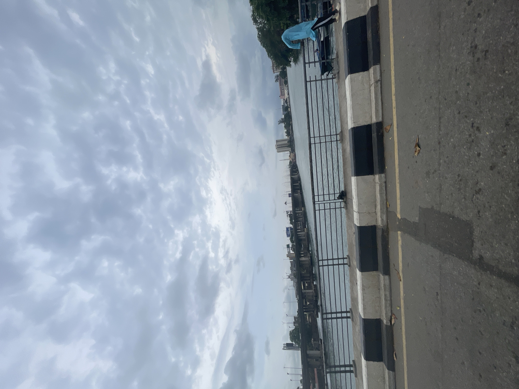 The scene shows a waterfront area in Lagos, Nigeria. A body of water is separated from a road by a black and white painted concrete barrier and a metal railing. The sky is overcast with grey clouds. The road surface is asphalt and shows signs of wear and discoloration. There's a garment draped over the railing and buildings in the distance across the water.