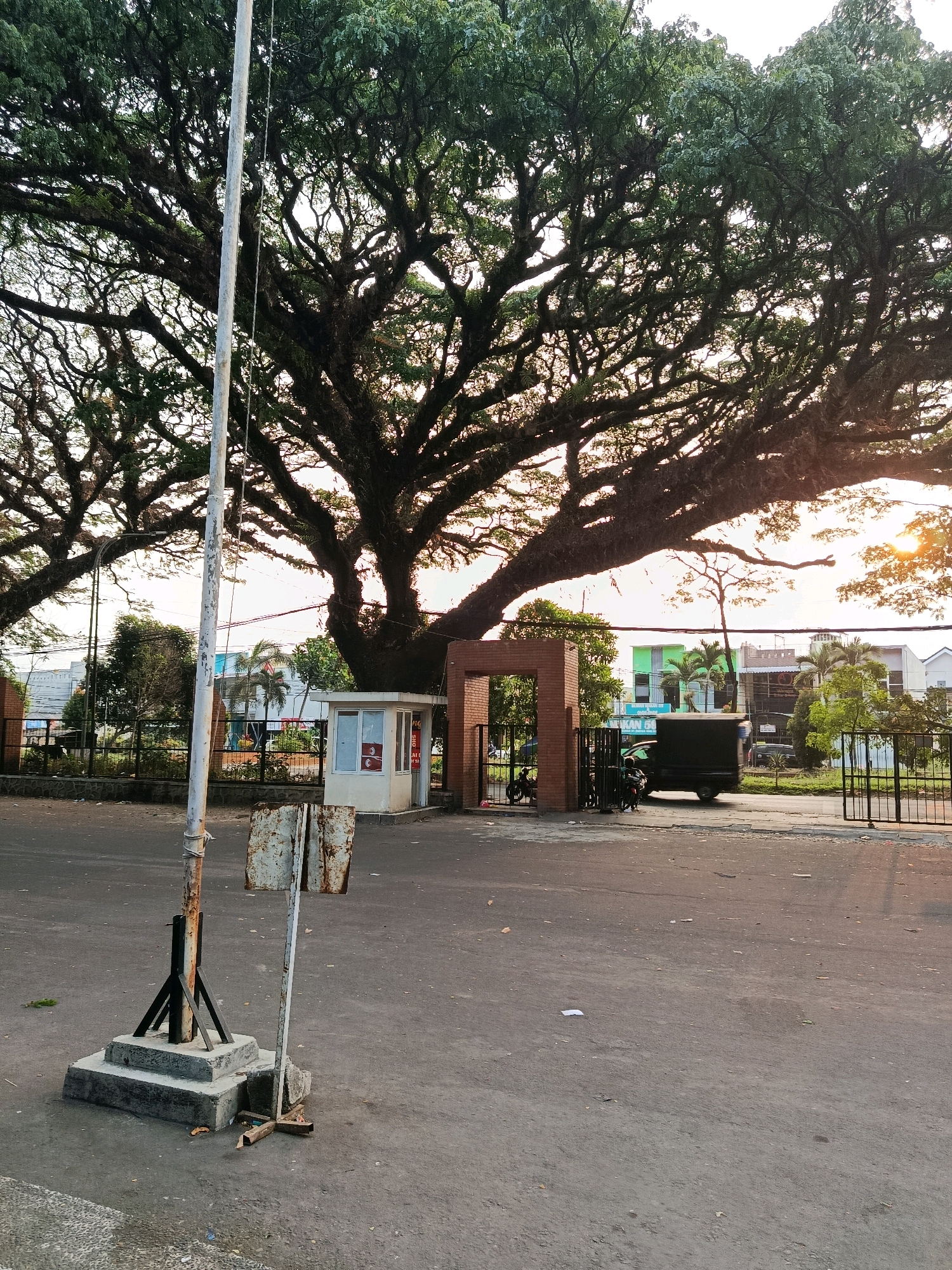 The image depicts a scene in the city of Malang, Indonesia, in front of a large, old tree. In the foreground, a weathered metal pole stands on a concrete base, with a rusty, discarded sign leaning against it. To the right, a small, white security booth with red signage is visible. Behind the booth, a brick archway leads to a fenced area. A black truck and two motorcycles are parked near the entrance. In the background, buildings and palm trees are seen under a bright, hazy sky, with the sun's rays filtering through the tree canopy. The ground is paved with asphalt and shows scattered debris.