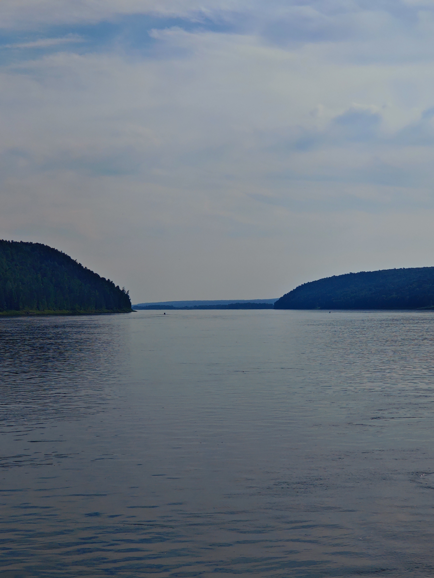 The image depicts a wide, calm river with a person in a small boat visible in the distance. On the left side of the river, a densely forested hill slopes down to the water. On the right side, a similar, less vegetated hill forms a concave shoreline. The sky is overcast with light clouds. This scene is likely the Angara River in Predivinsk, Russia, a region known for its vast waterways and forested landscapes.